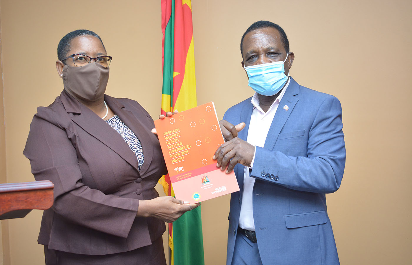 Elaine McQueen in brown suit presents publication to Prime Minister, Dr. Keith Mitchell who is wearing a blue suite. Grenada's flag is int he background