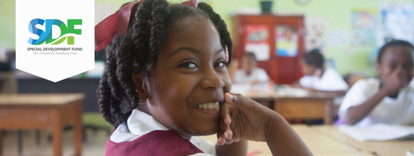 Black primary school girl with natural hair in twists and dressed in maroon uniform with white inside shirt seated in a classroom looking over her shoulder into the camera