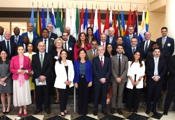group photo of males and females of various ethnicities in business attire standing in front a row of country flags