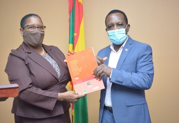 Elaine McQueen in brown suit and mask presents report to Prime Minister, Dr. Keith Mitchell who is wearing a blue suit and mask. The Grenada flag is in the background.