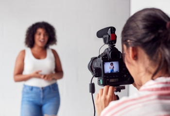 Woman with camera recording female vlogger in white top and blue jeans.