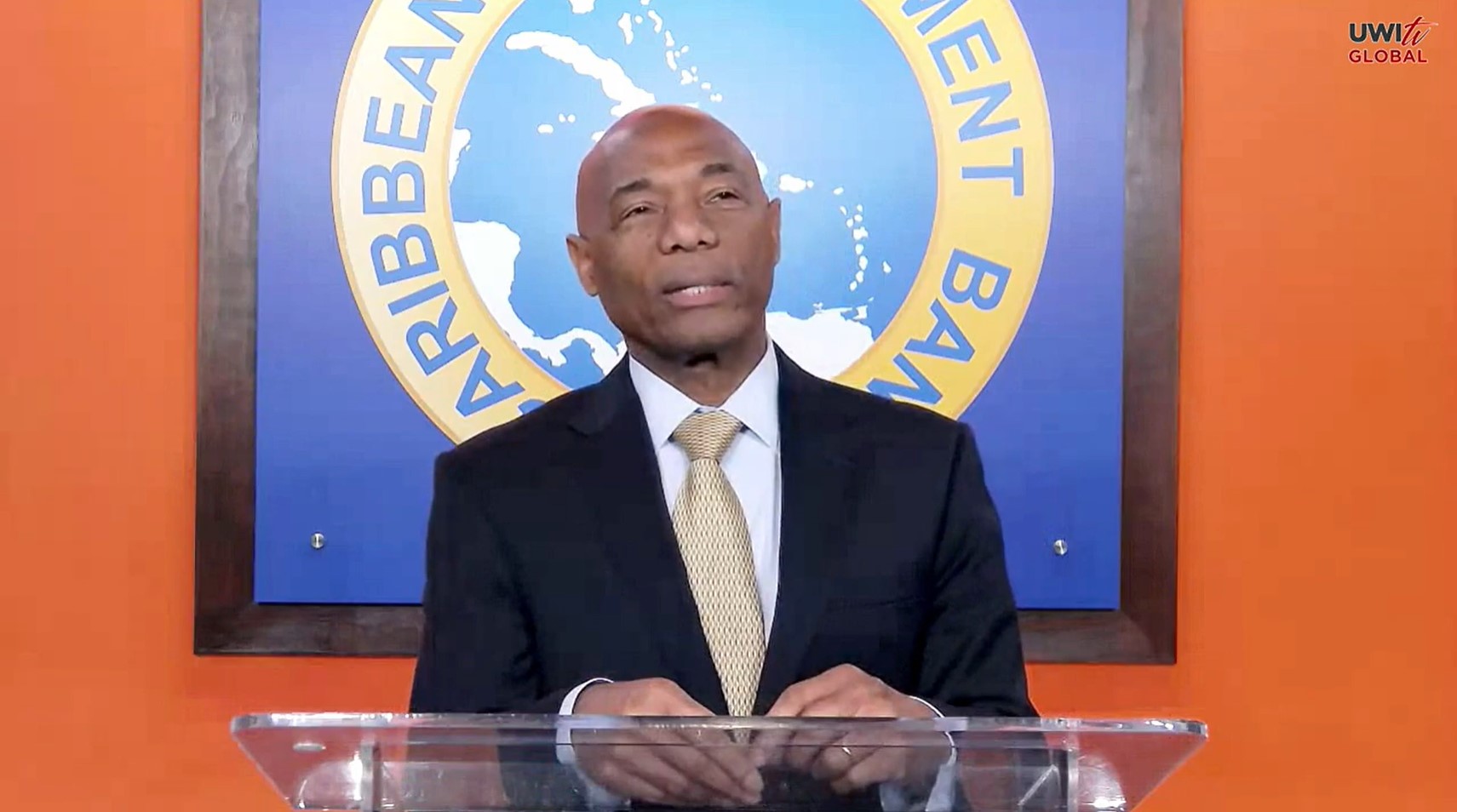 CDB President Dr Gene Leon at lectern with orange background and large CDB logo behind him.