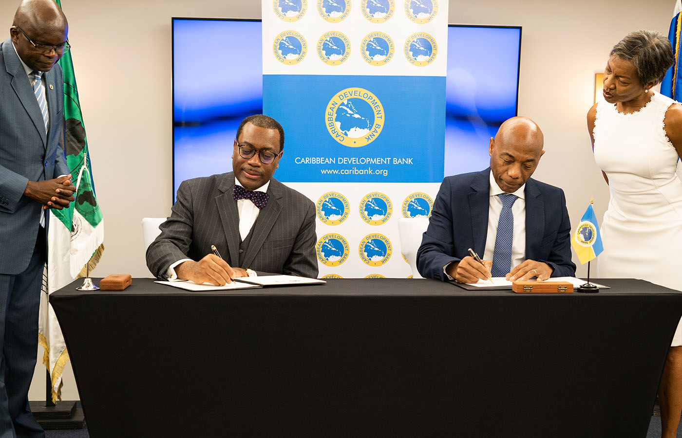 two seated gentlemen signing document as others look on