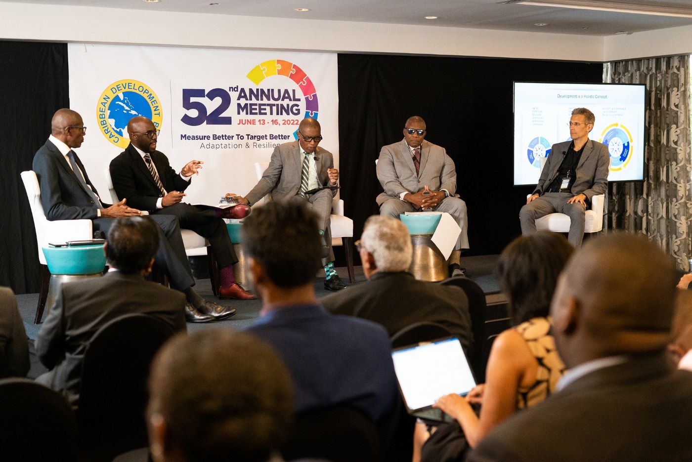 Five panellists seated on dark riser with multicoloured logo on screen behind them.