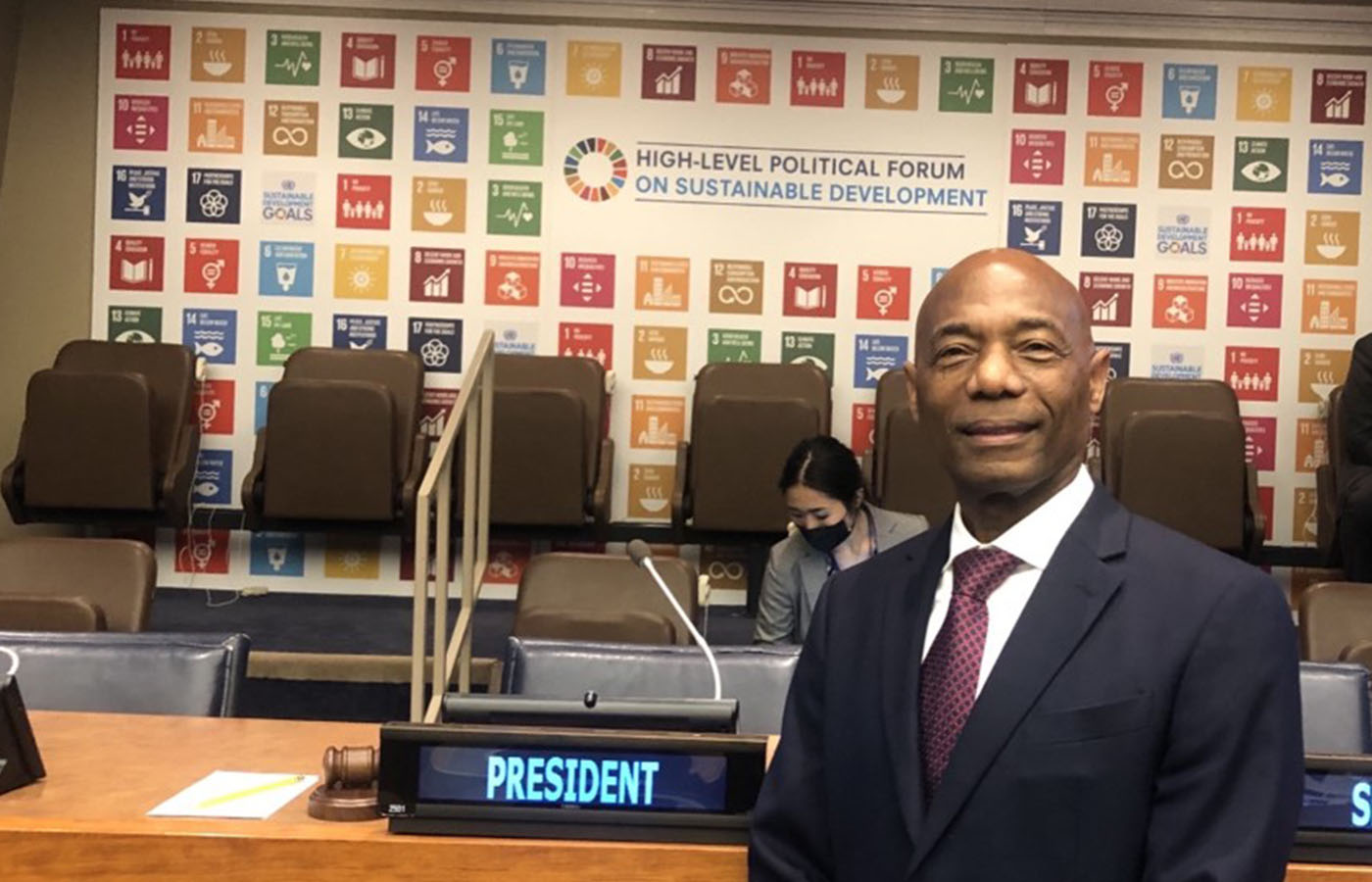 gentleman in dark suit with white shirt and red tie standing in front of a backdrop with the SDG icons