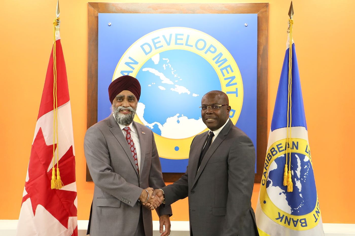 Minister Harjit Sajjan shaking hands with CDB Vice President Isaac Solomon in front of flags of Canada and CDB