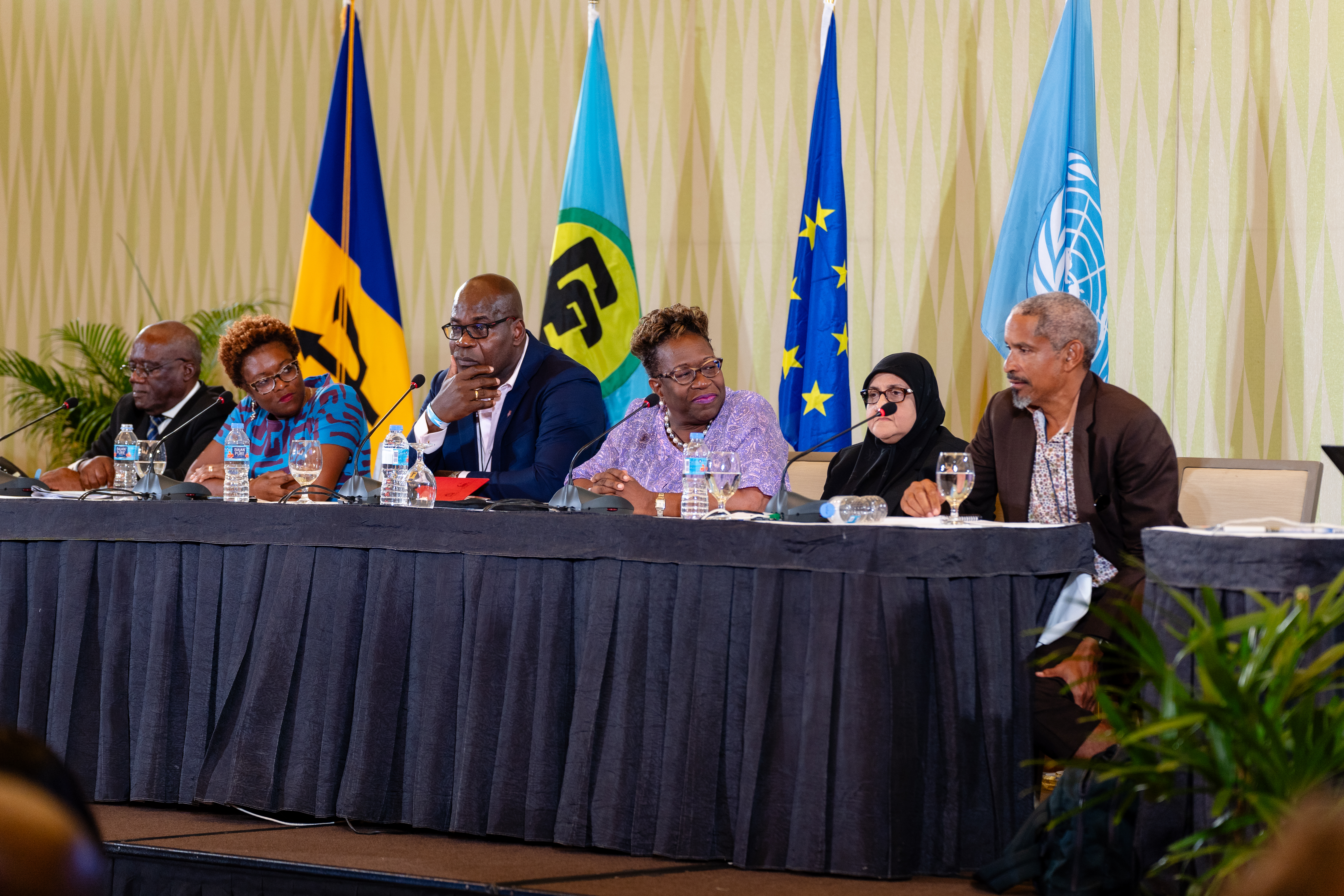 Six persons sit at the head table with the flags of Barbados, CARICOM, the EU and United Nations behind them 