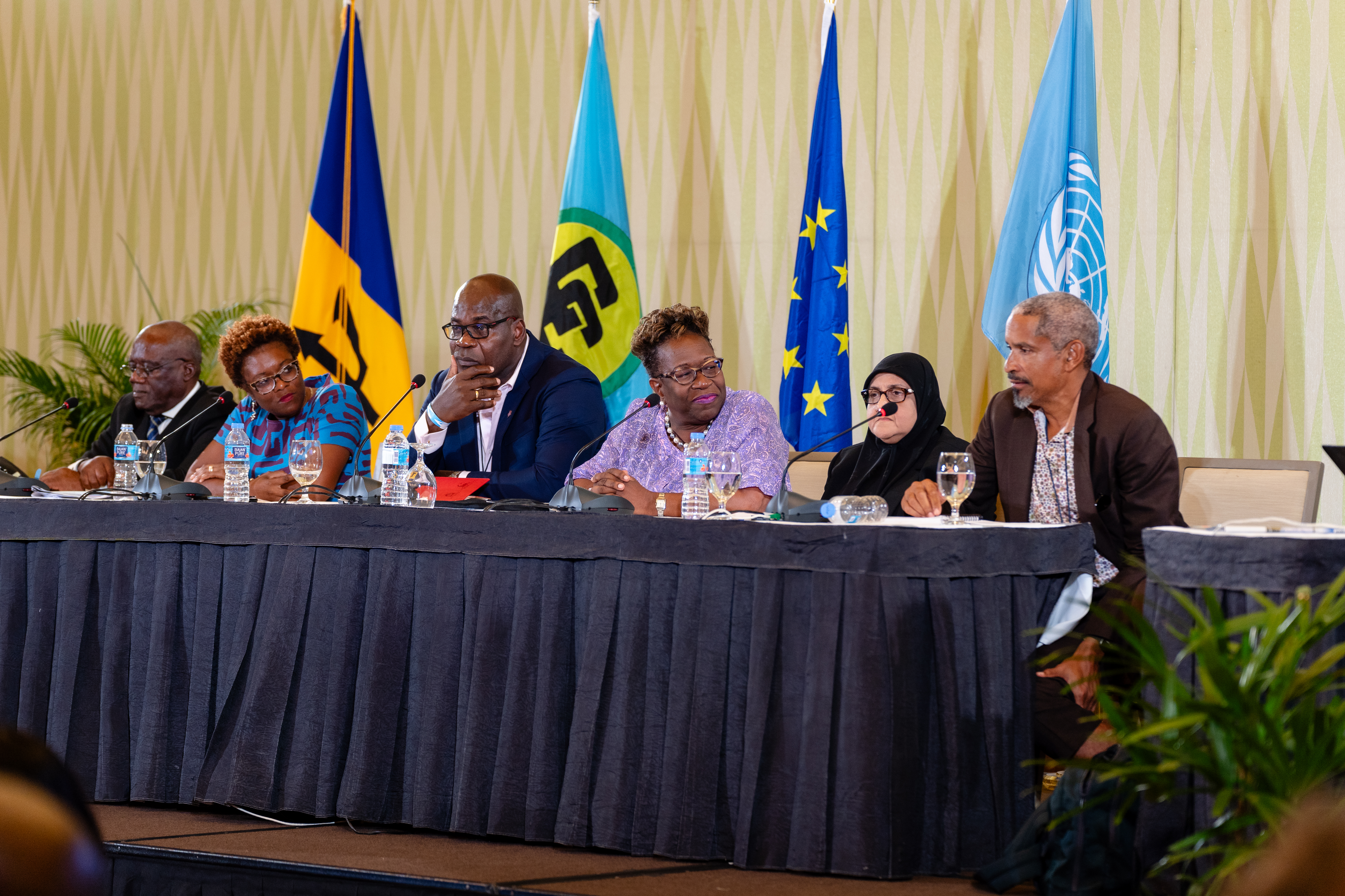 Six persons sit at the head table with the flags of Barbados, CARICOM, the EU and United Nations behind them