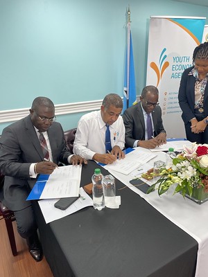 Three men are sitting at a table signing a document with a woman standing watching as they sign