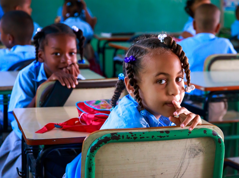 Two young girls in school uniform in a classroom have turned around staring at the camera 