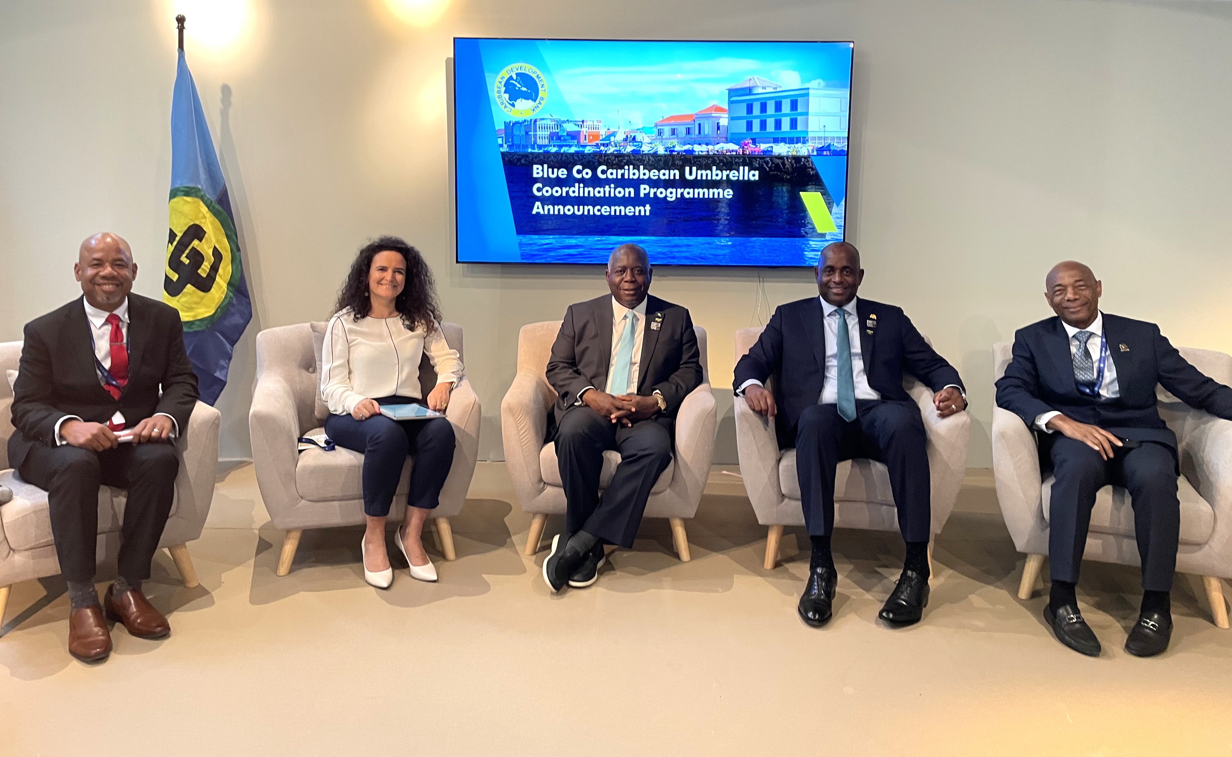 Five individuals are seated in a room below a television screen with a CARICOM flag to the right 