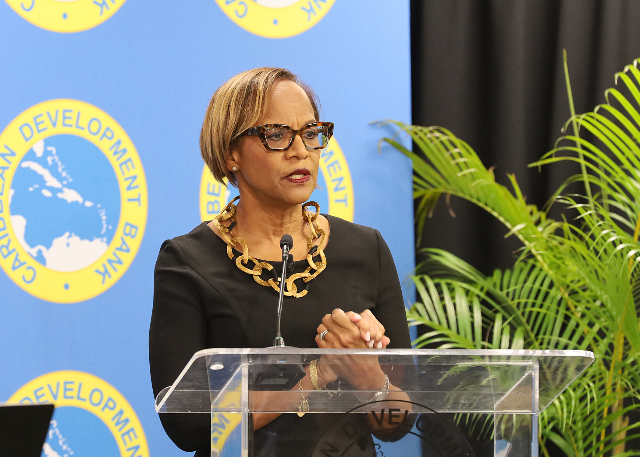 black woman waring glasses and a black dress standing at acrylic lectern with step and repeat backdrop behind her