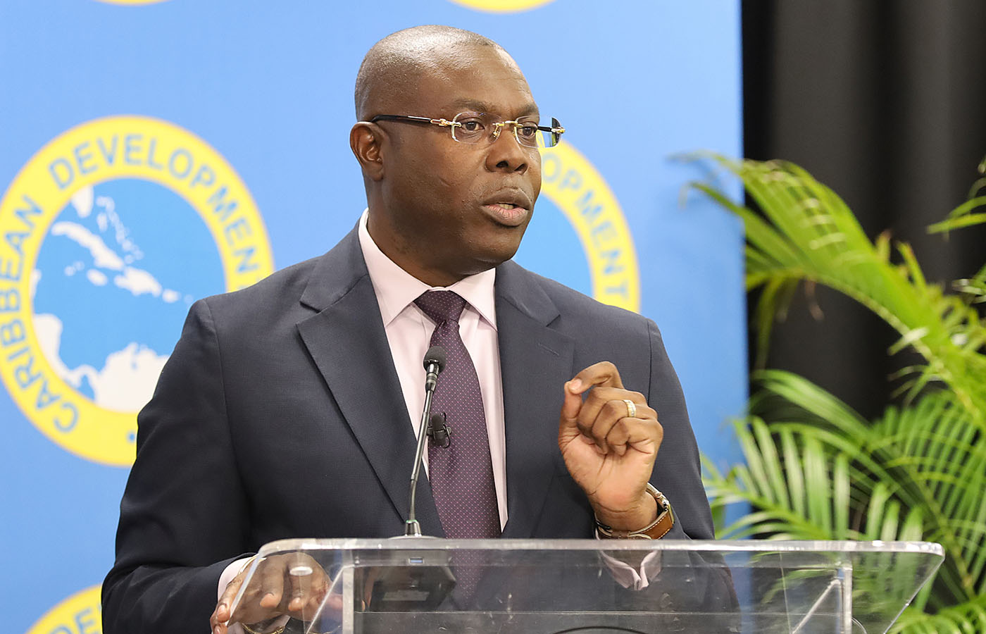 black man wearing glasses in dark suit with pink shirt and mauve tie standards at acrylic lectern delivering remarks