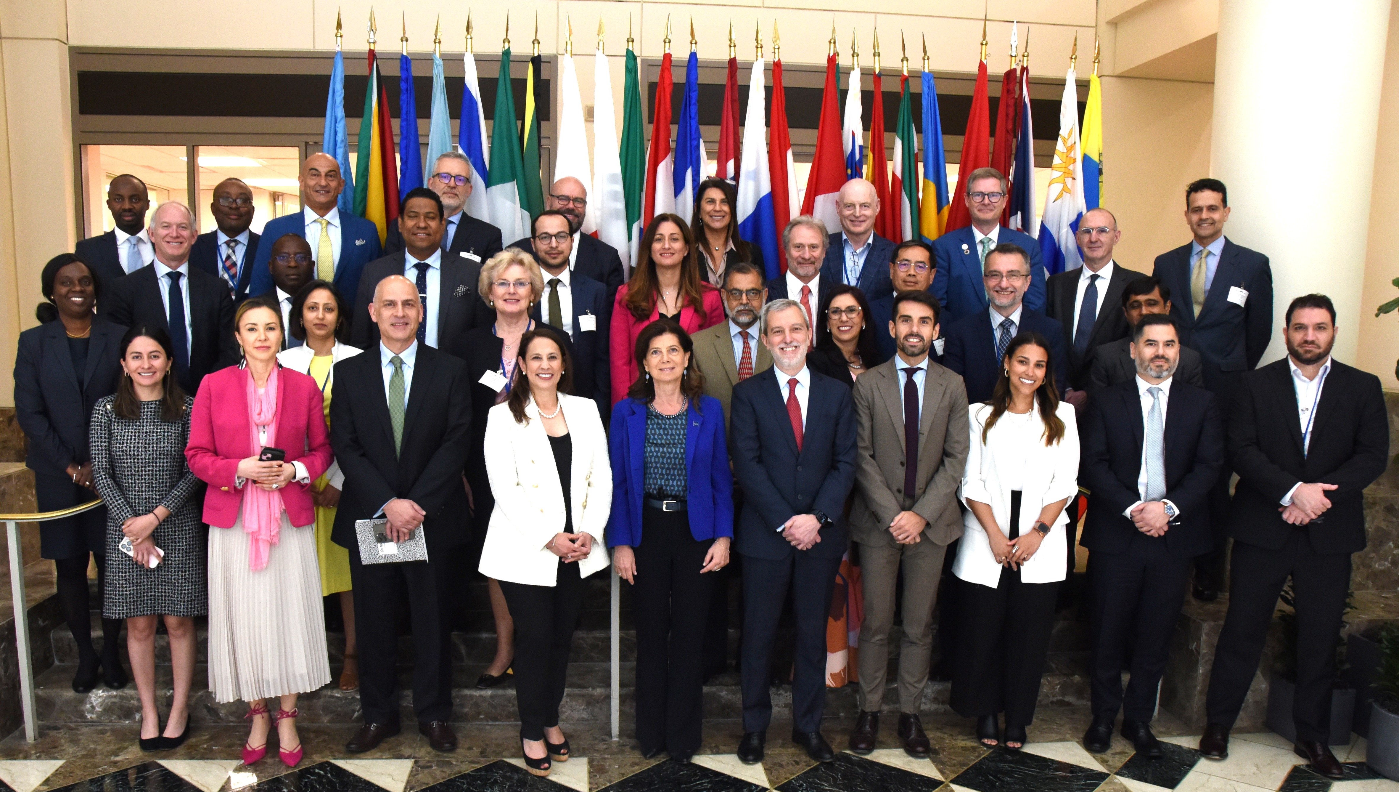 group photo of males and females of various ethnicities in business attire standing in front a row of country flags