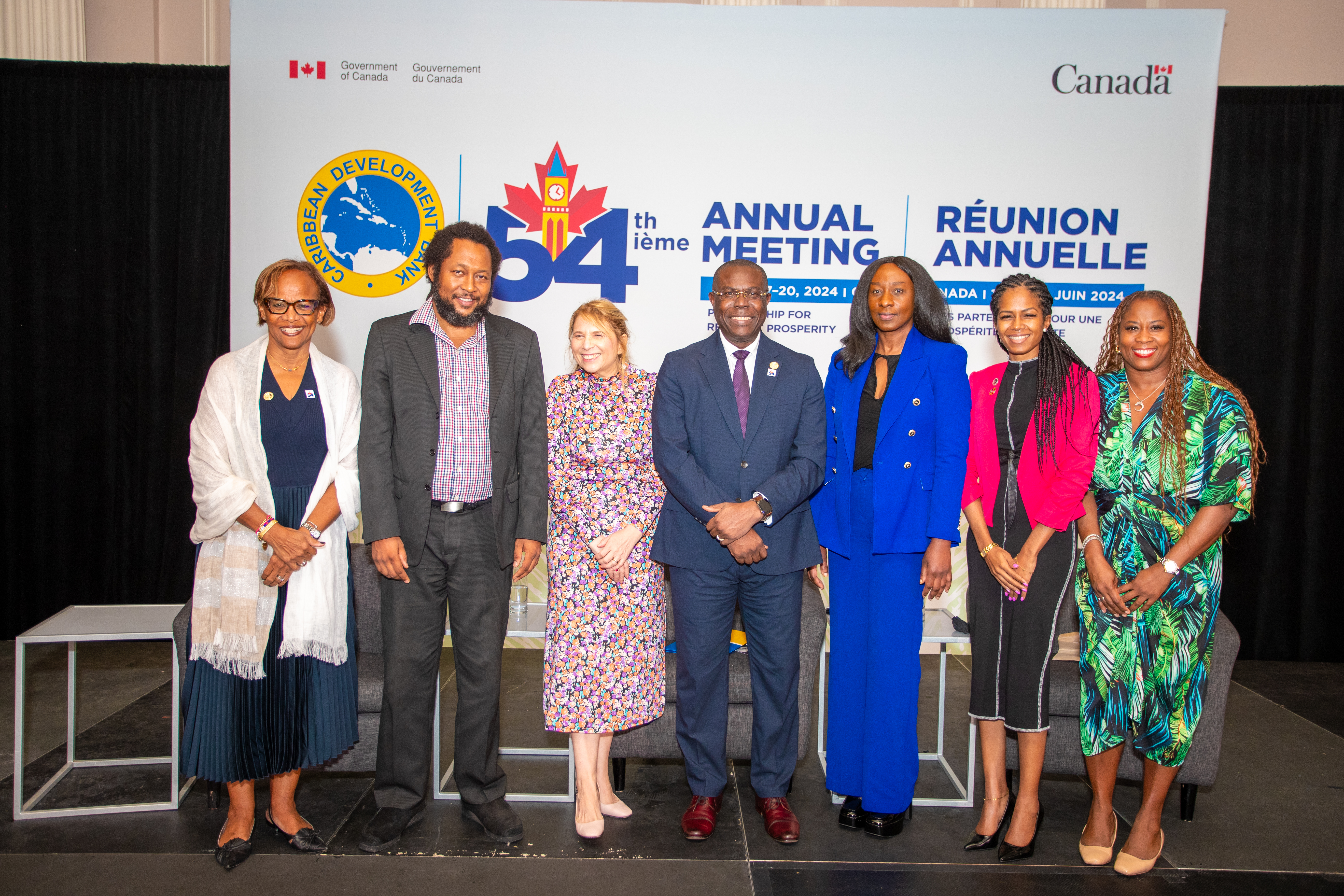 seven people are standing alongside each other on a platform gainst the CDB 54th Annual Meeting backdrop