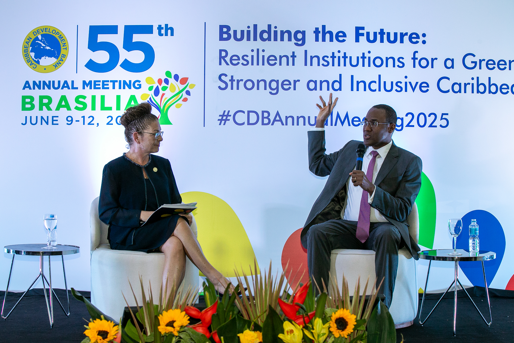 Two individuals are seated on a stage at the 55th Annual Meeting of the Caribbean Development Bank in Brasília, Brazil. On the left, a woman holds a notebook, attentively listening, while on the right, a man gestures while speaking into a microphone. A colorful backdrop displays the event's theme, "Building the Future: Resilient Institutions for a Greener, Stronger, and Inclusive Caribbean."