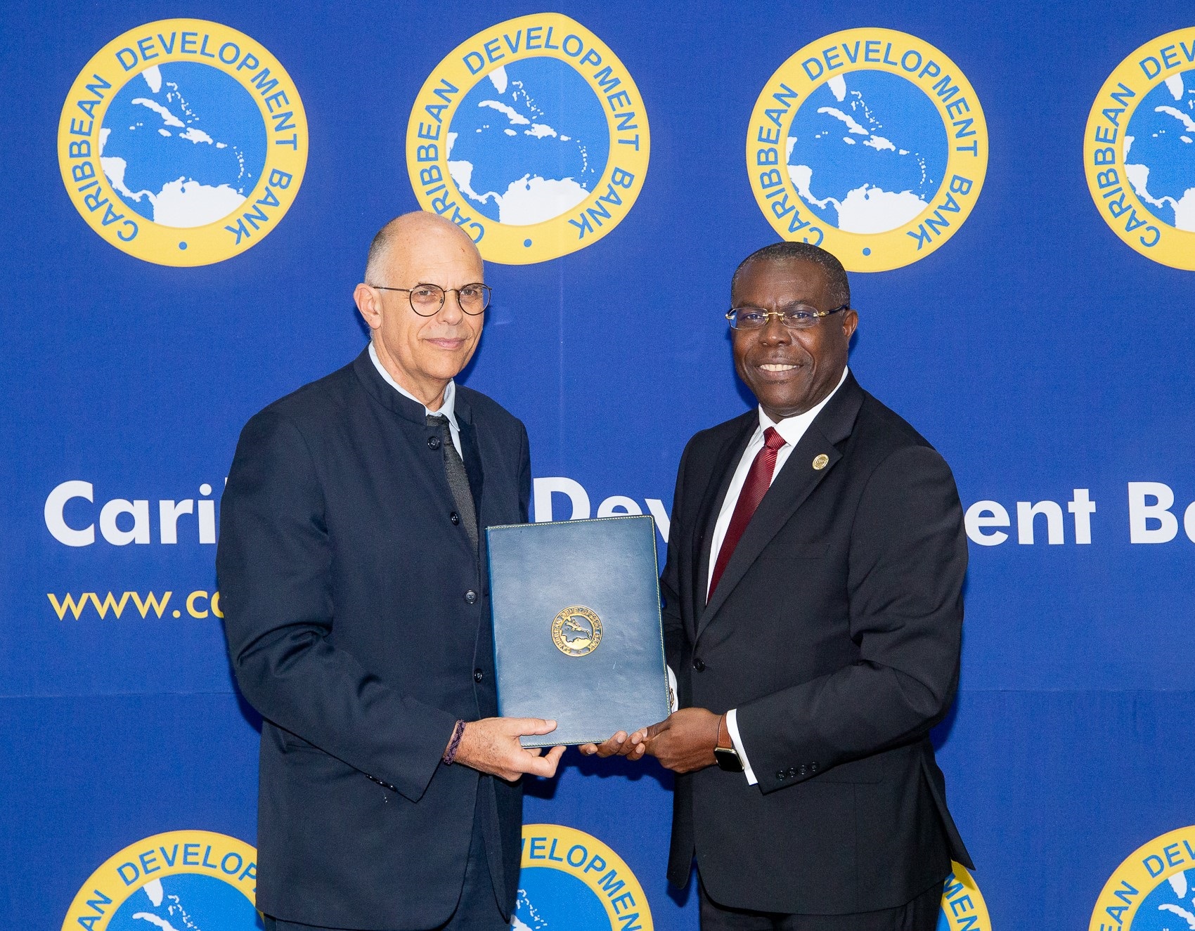 Two men in formal attire standing front of a blue backprop with the Caribbean Development Bank logo. They are smiling and holding a blue folder embossed with the Bank’s seal, during the signing ceremony.