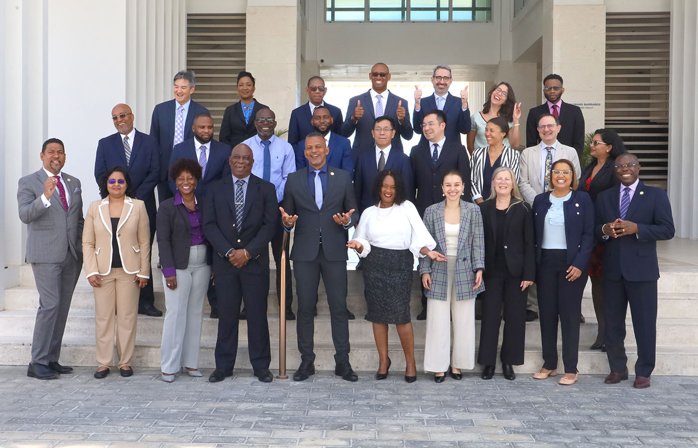 A diverse group of professionals stands together on the steps outside a modern building for a group photo. The individuals are smiling, gesturing positively, and dressed in formal business attire, indicating a celebratory or official event. The setting suggests a conference or institutional gathering, with architectural columns and large windows in the background.