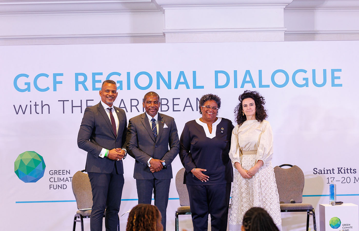 A photo from the GCF Regional Dialogue with the Caribbean, held in Saint Kitts and Nevis from March 17–20, 2025. Four people are standing on stage in front of a banner displaying the event name and Green Climate Fund logo. From left to right: a man in a dark suit, a man in a navy suit, a woman in a navy pantsuit with a pin, and a woman in a cream dress. 
