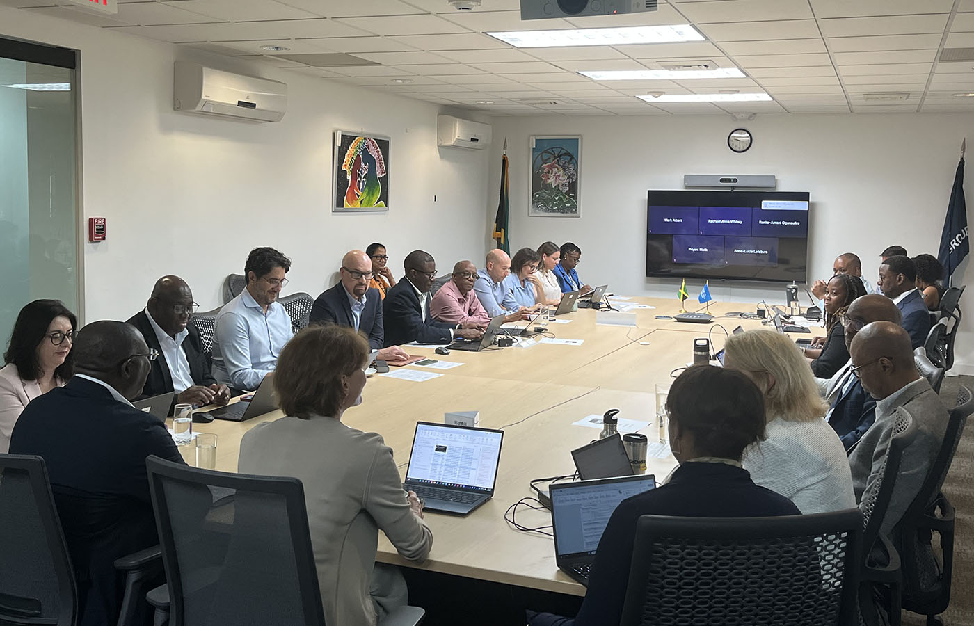 A group of professionals from the Caribbean Development Bank and the World Bank are seated around a large rectangular conference table in a modern meeting room. Most participants are using laptops, and several are engaged in discussion. On the wall ahead is a large screen displaying names of remote attendees, indicating a hybrid meeting setup. Flag of Jamaica is in the left corner.