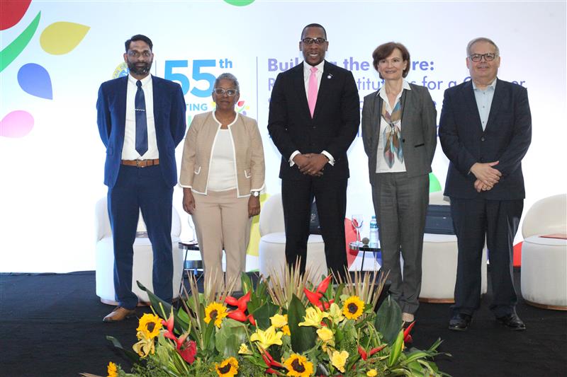 **Description for Accessibility:** A group of five individuals stands on a stage at the 55th Annual Meeting of the Caribbean Development Bank, with the event's theme "Building the Future: Resilient Institutions for a Greener, Stronger, and Inclusive Caribbean" displayed on the backdrop. The individuals, representing a mix of formal attire, are positioned in front of a vibrant floral arrangement featuring sunflowers and tropical plants. The event logo is prominently visible in the background.