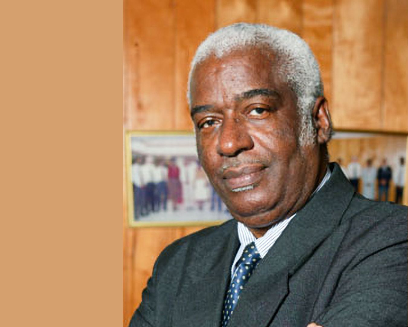 Corporate portrait of a grey haired elderly black man in a black business suit against a brown background
