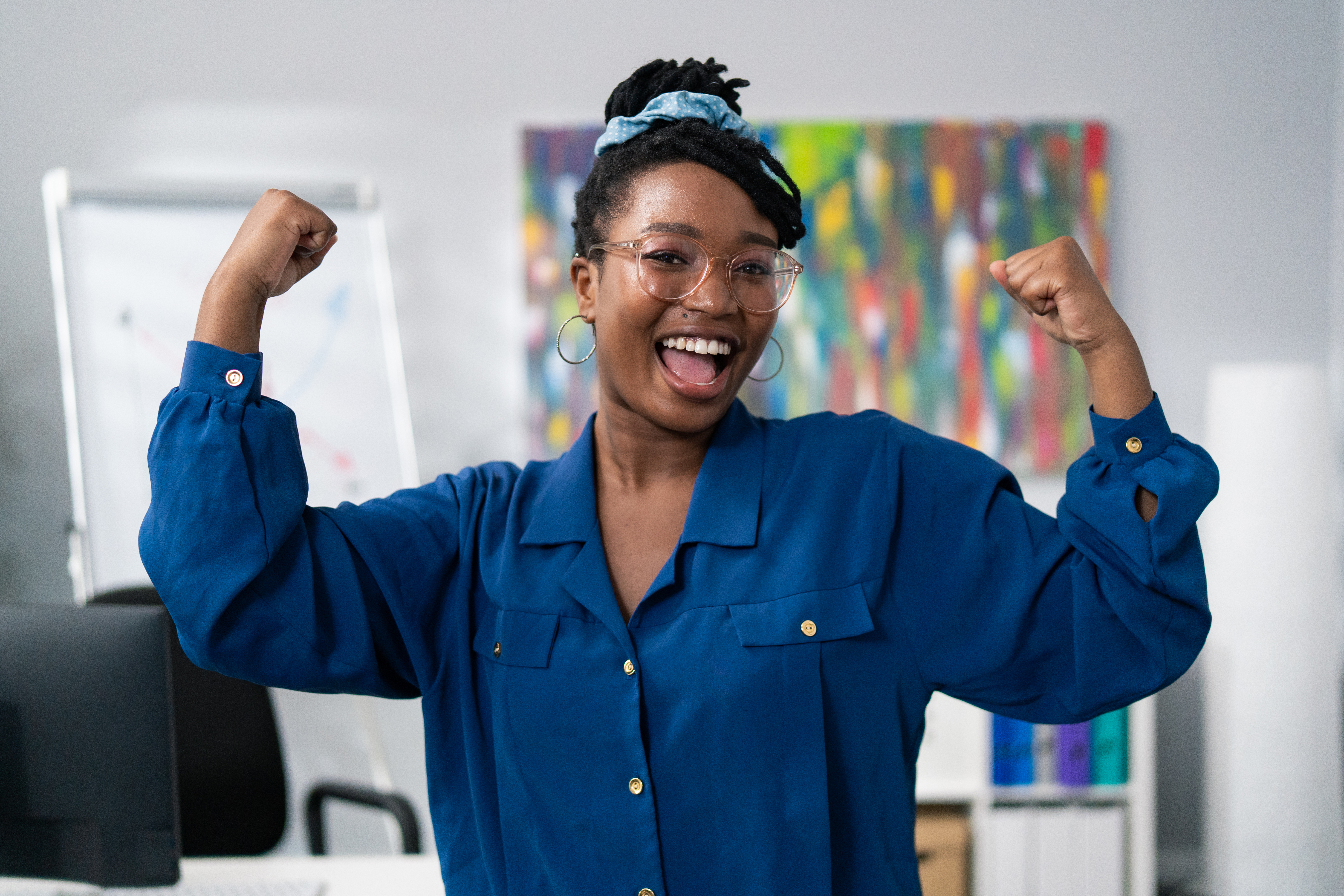 Dark-skinned woman wearing blue clothes glasses stands against office backdrop smiles happy, victory, winning, raises hands up tenses hand muscles gestures strength power successful girl businesswoman