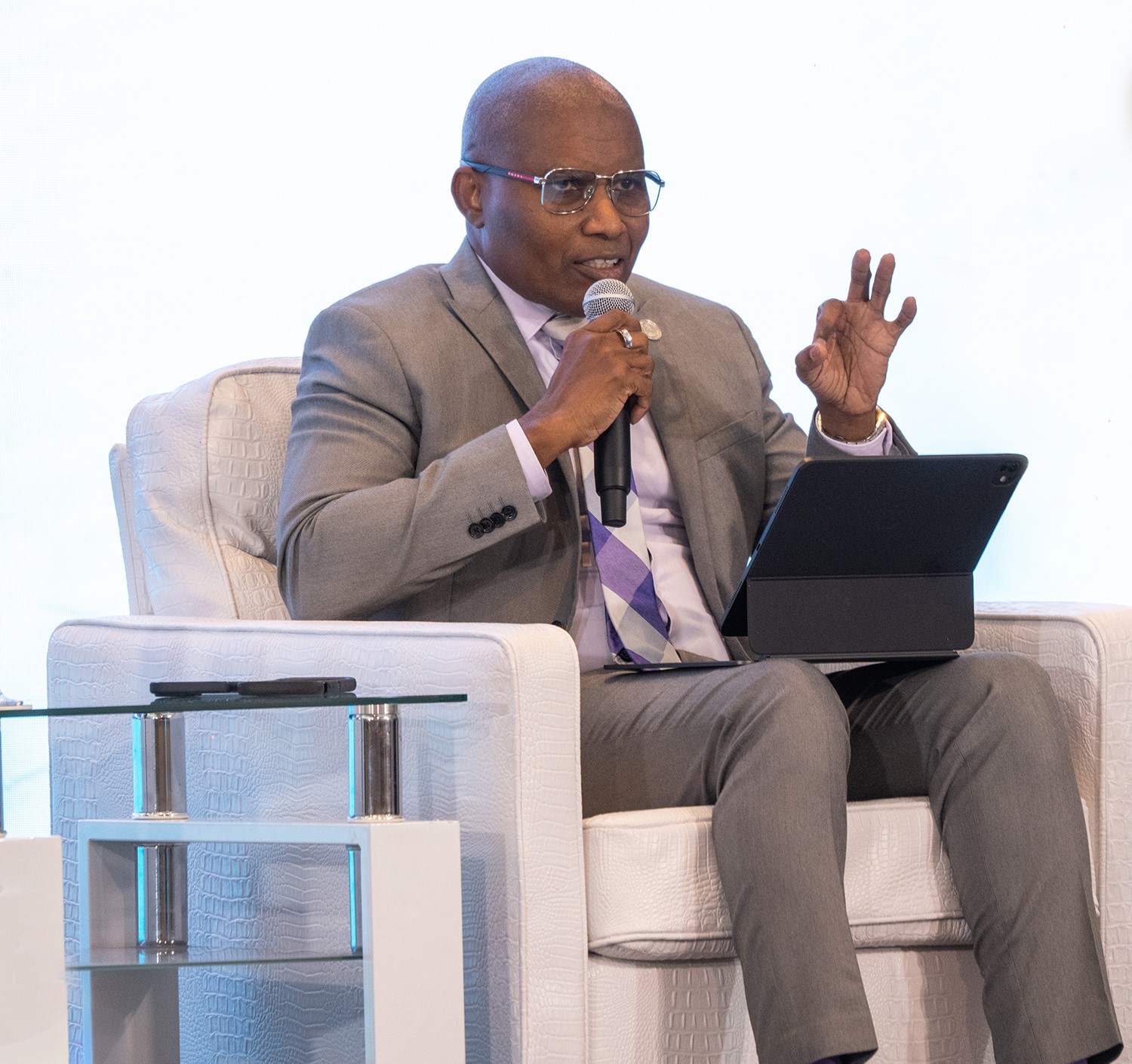 Bespectacled black man seated in a grey suit on a white chair on stage with a mic in hand gesticulating and looking ahead 