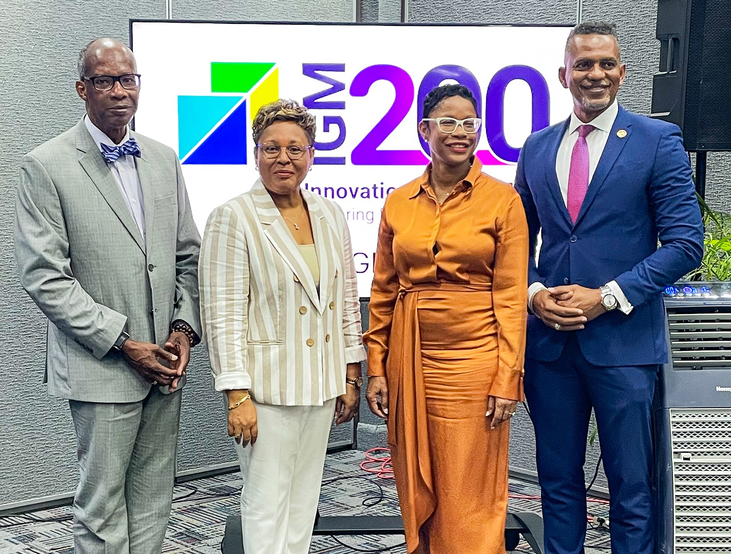 a group of four black persons two males one in a grey suit and another in a blue suit flanking two ladies one in white and one in orange all standing looking ahead 