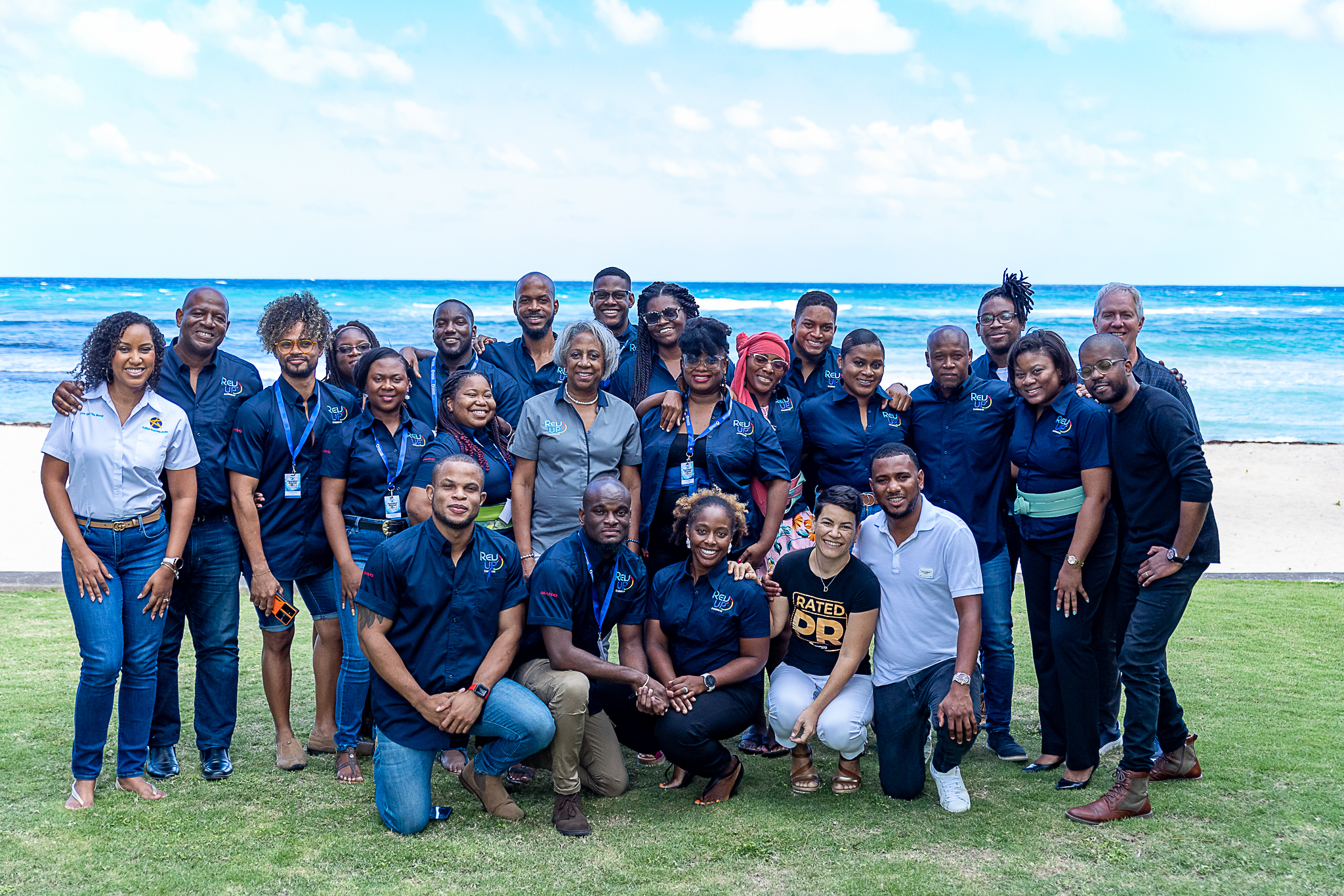 A large group of black entrepreneurs standing and stooping in navy blue tshirts on grass under a clear blue sky and ocean