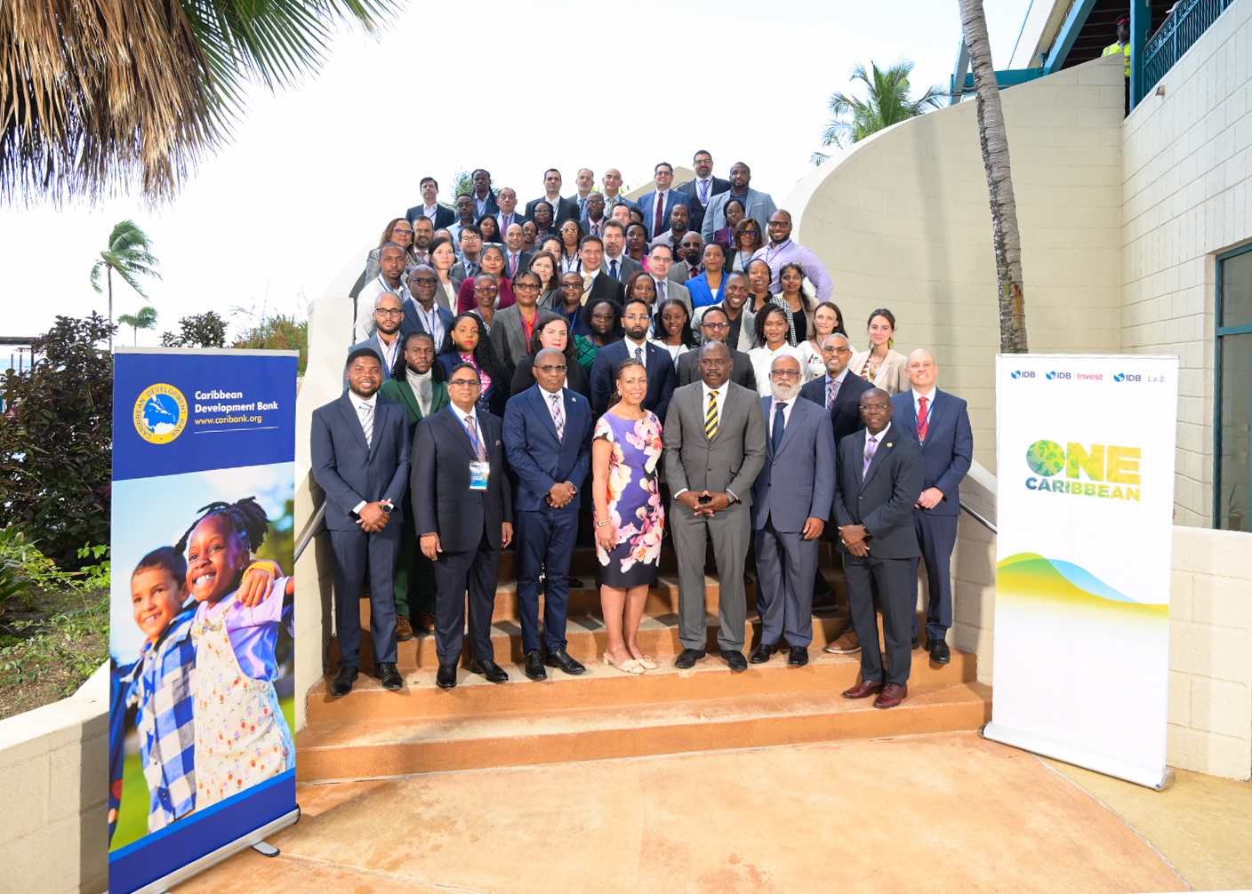 Large group of delegates standing on stairs outdoors in business attire flanked by two banners