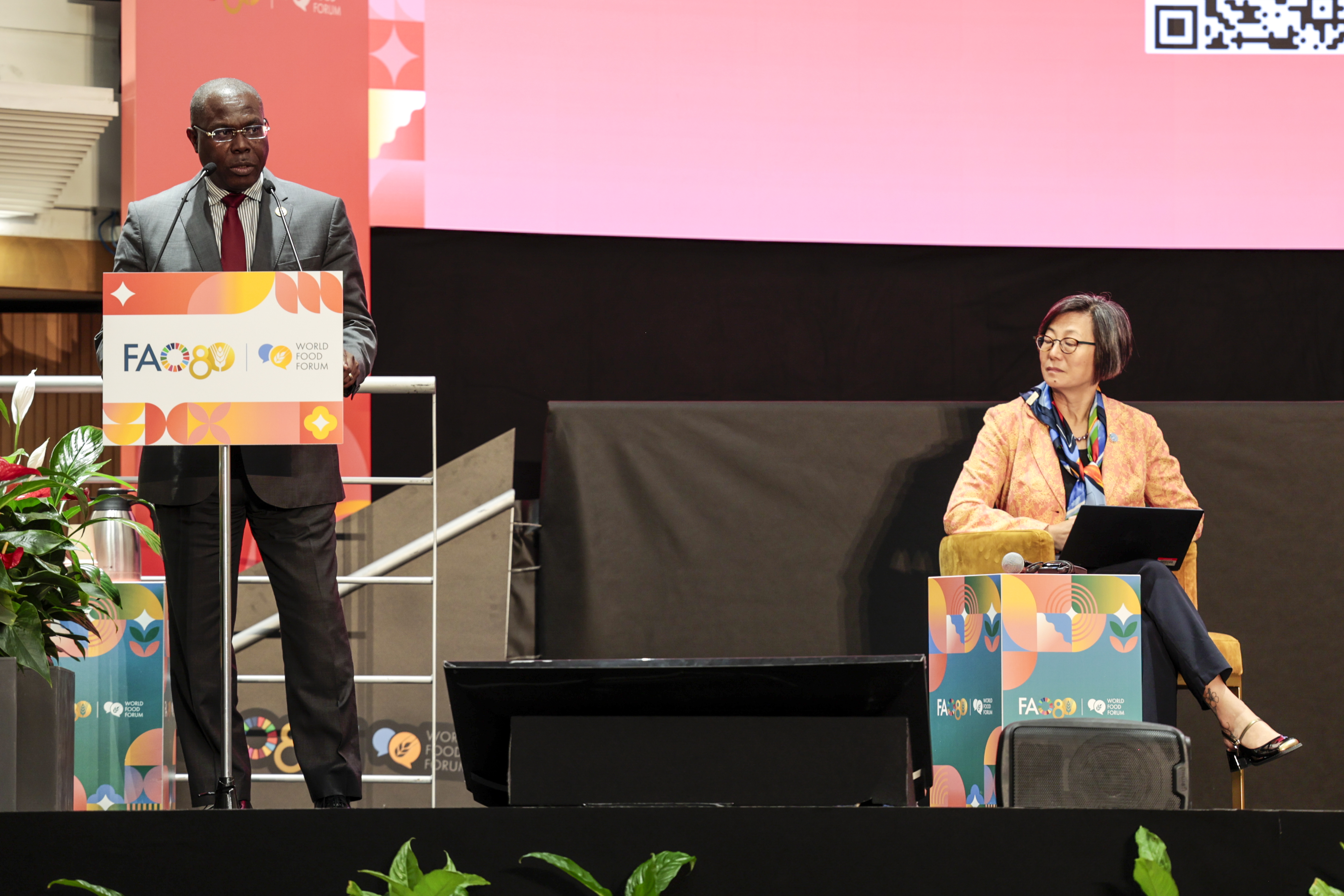 Isaac Solomon standing at lectern on stage on business attire with seated colleague 