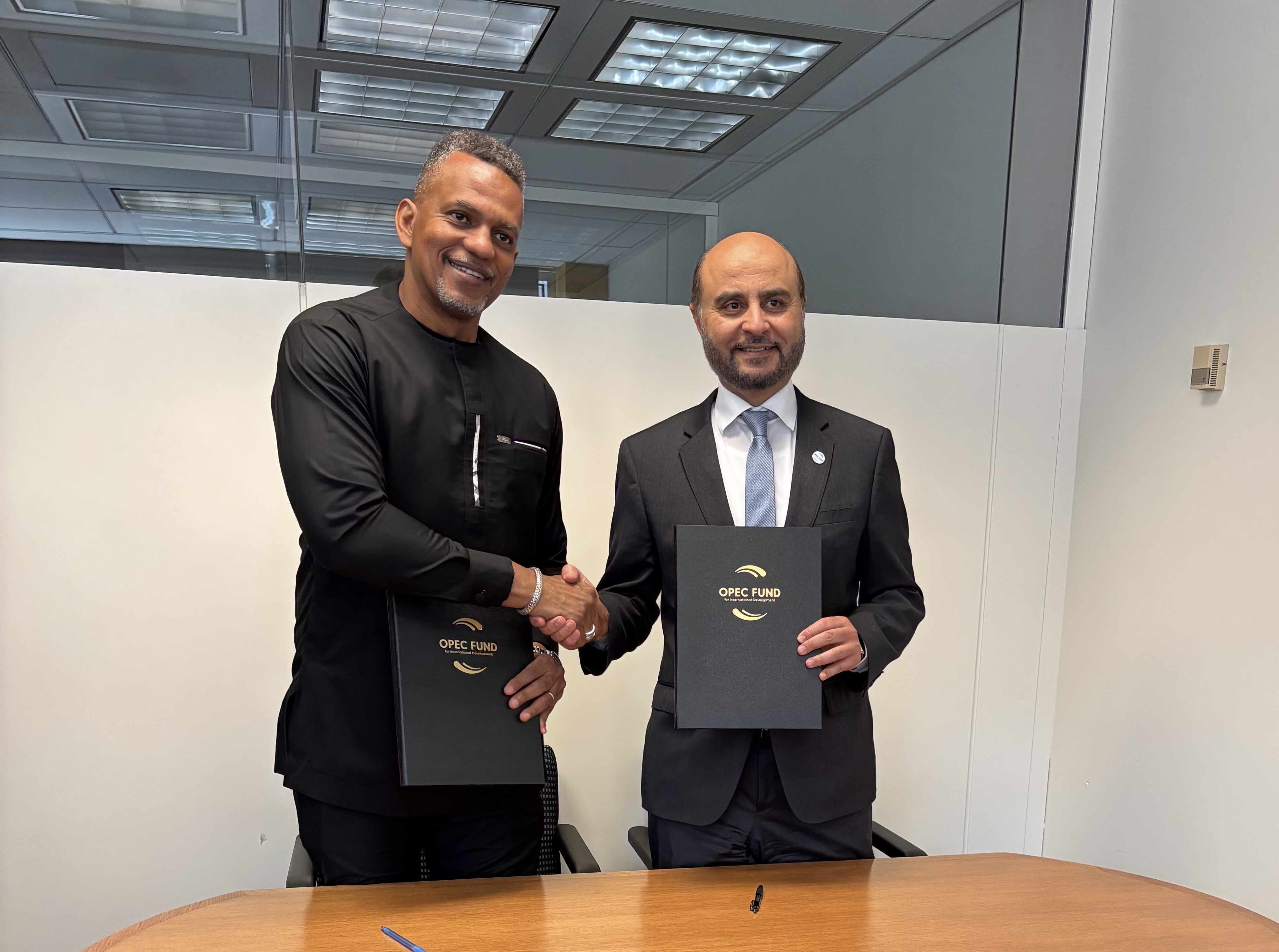 two men in business attire standing in a room behind a table holding up branded folders and shaking hands