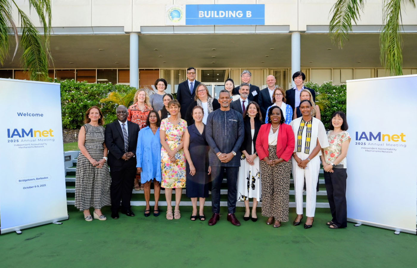 Group photo of participants at the IAMnet 2025 Annual Meeting in Bridgetown, Barbados, standing in front of Building B with event banners on both sides.