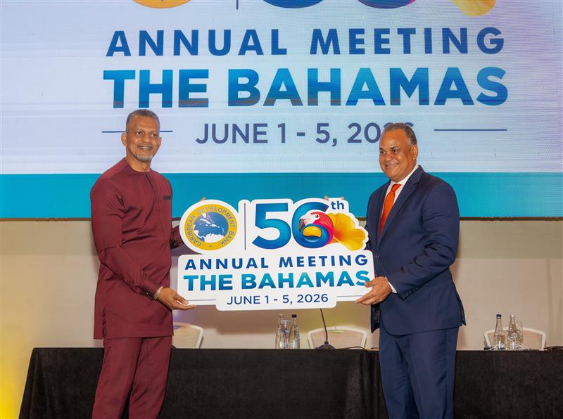 Chairman Halkitis and President Best holding a sign for the Caribbean Development Bank’s 56th Annual Meeting in The Bahamas, with event dates displayed on a stage.
