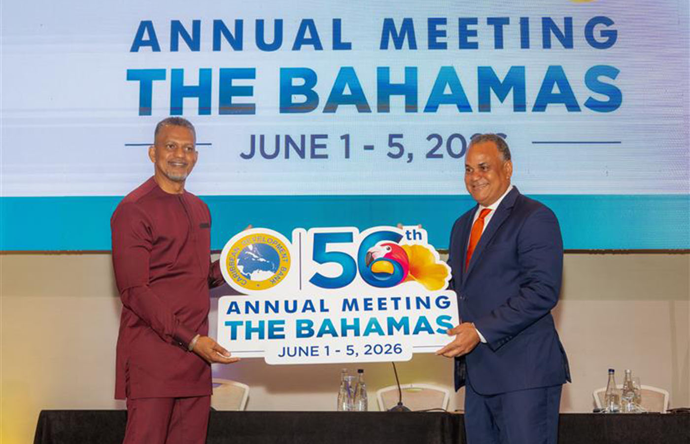 Chairman Halkitis and President Best holding a sign for the Caribbean Development Bank’s 56th Annual Meeting in The Bahamas, with event dates displayed on a stage.
