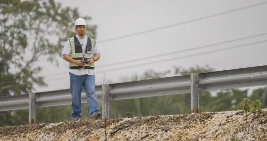 A man in a hard hat and ves walks along a highway.