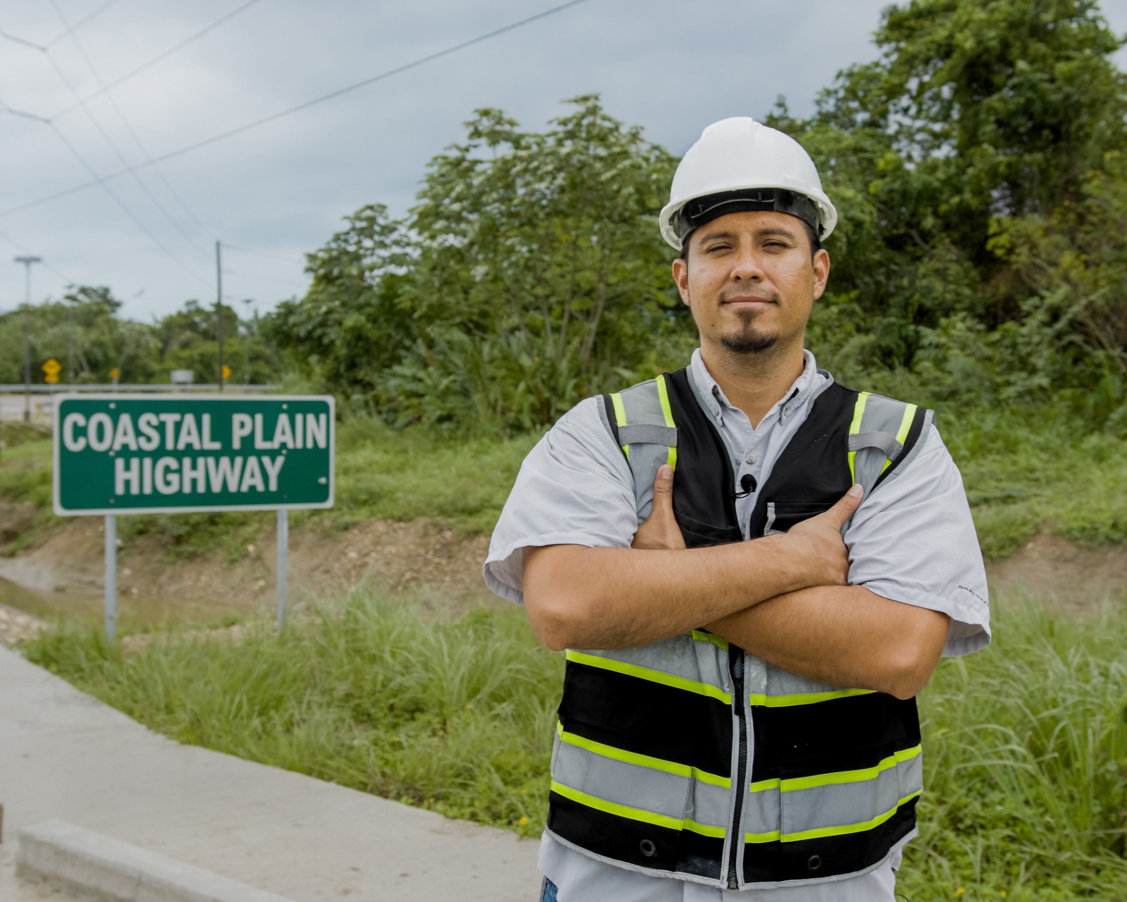 A man in a hard hat and vest stands with arms crossed on the shoulder of a highway.