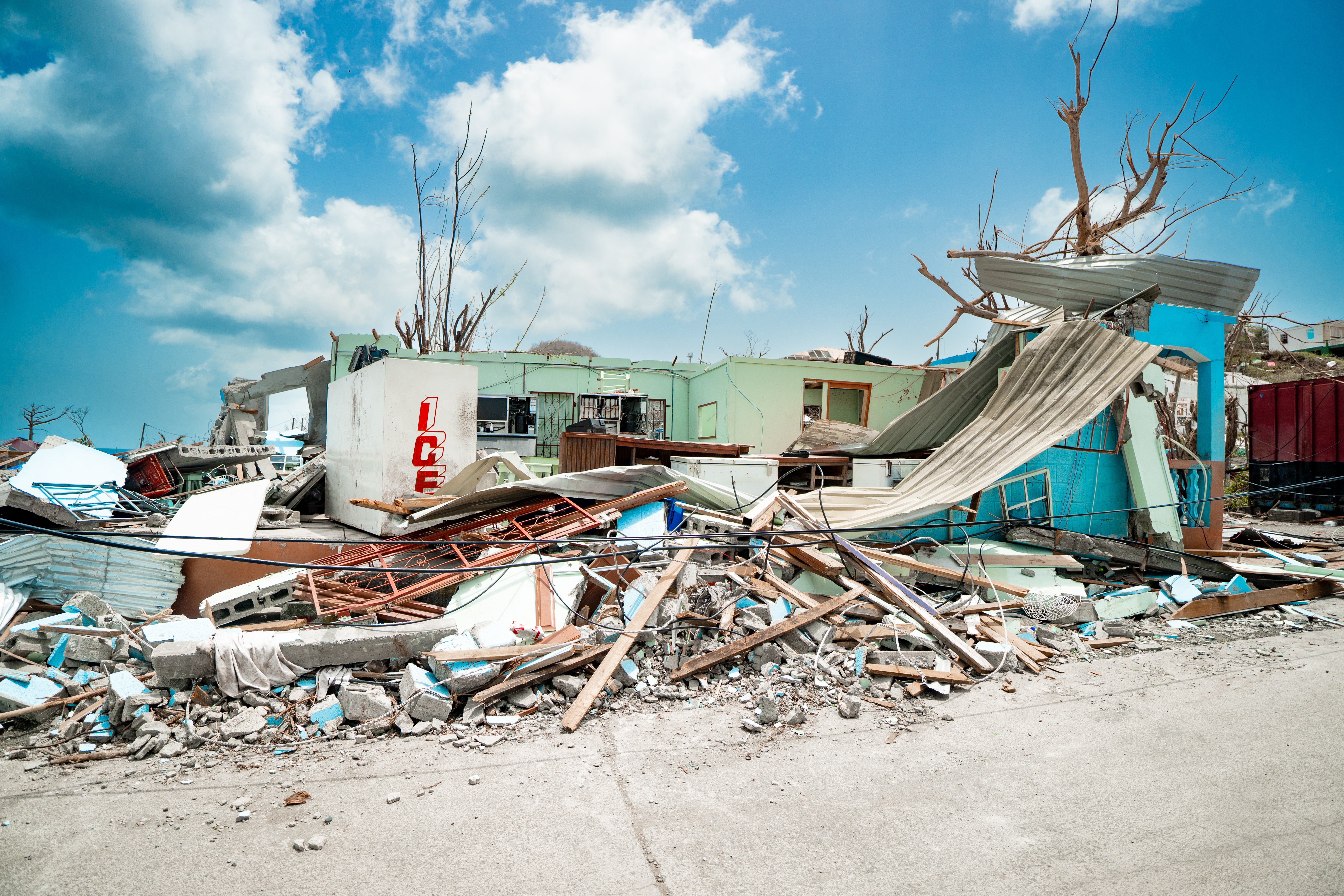Severely damaged buildings and debris scattered across a coastal area after a storm, with collapsed roofs and broken structures under a partly cloudy sky.