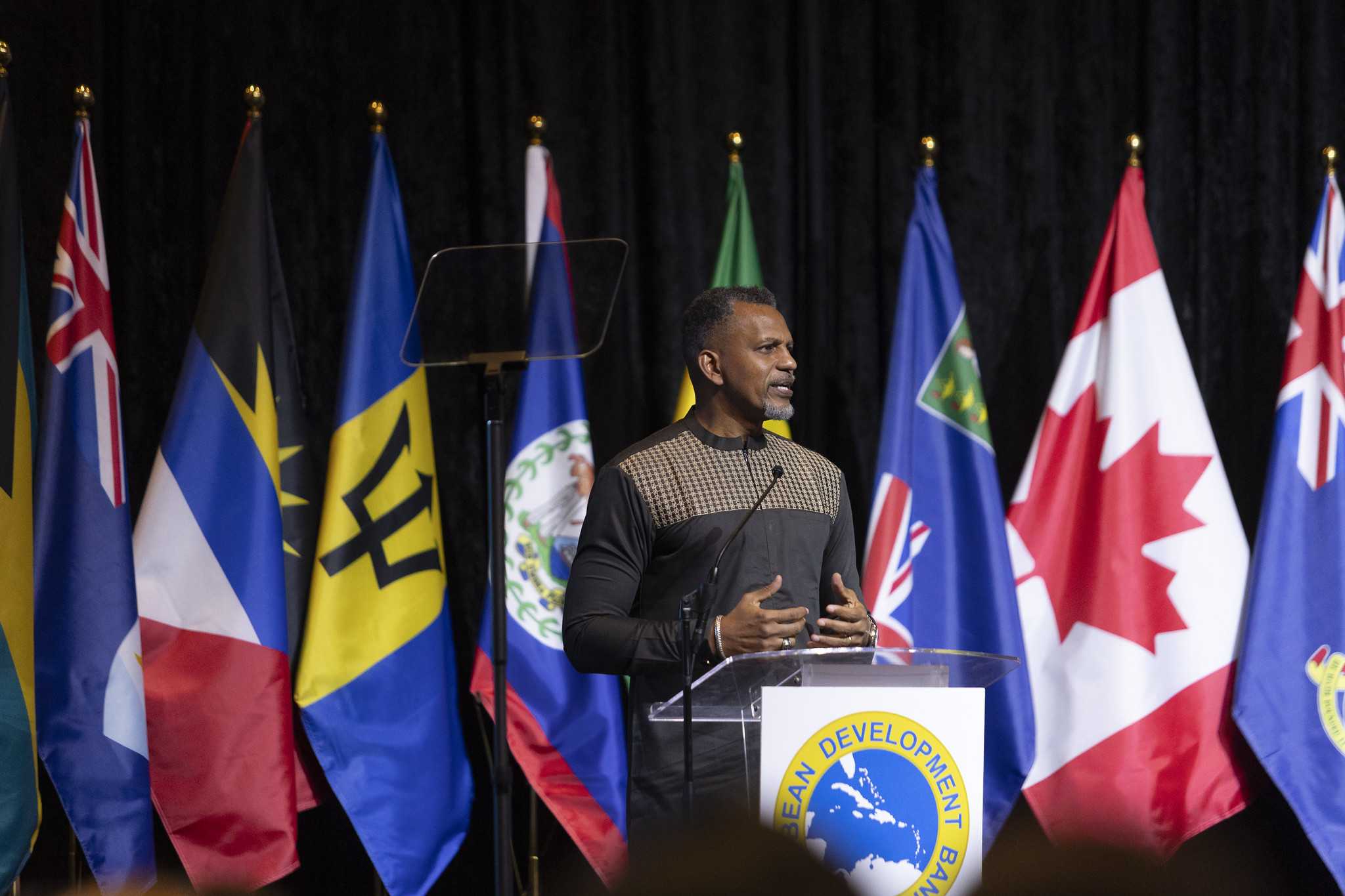 President of the Caribbean Development Bank delivers remarks from a podium on stage, with multiple national flags displayed behind him.