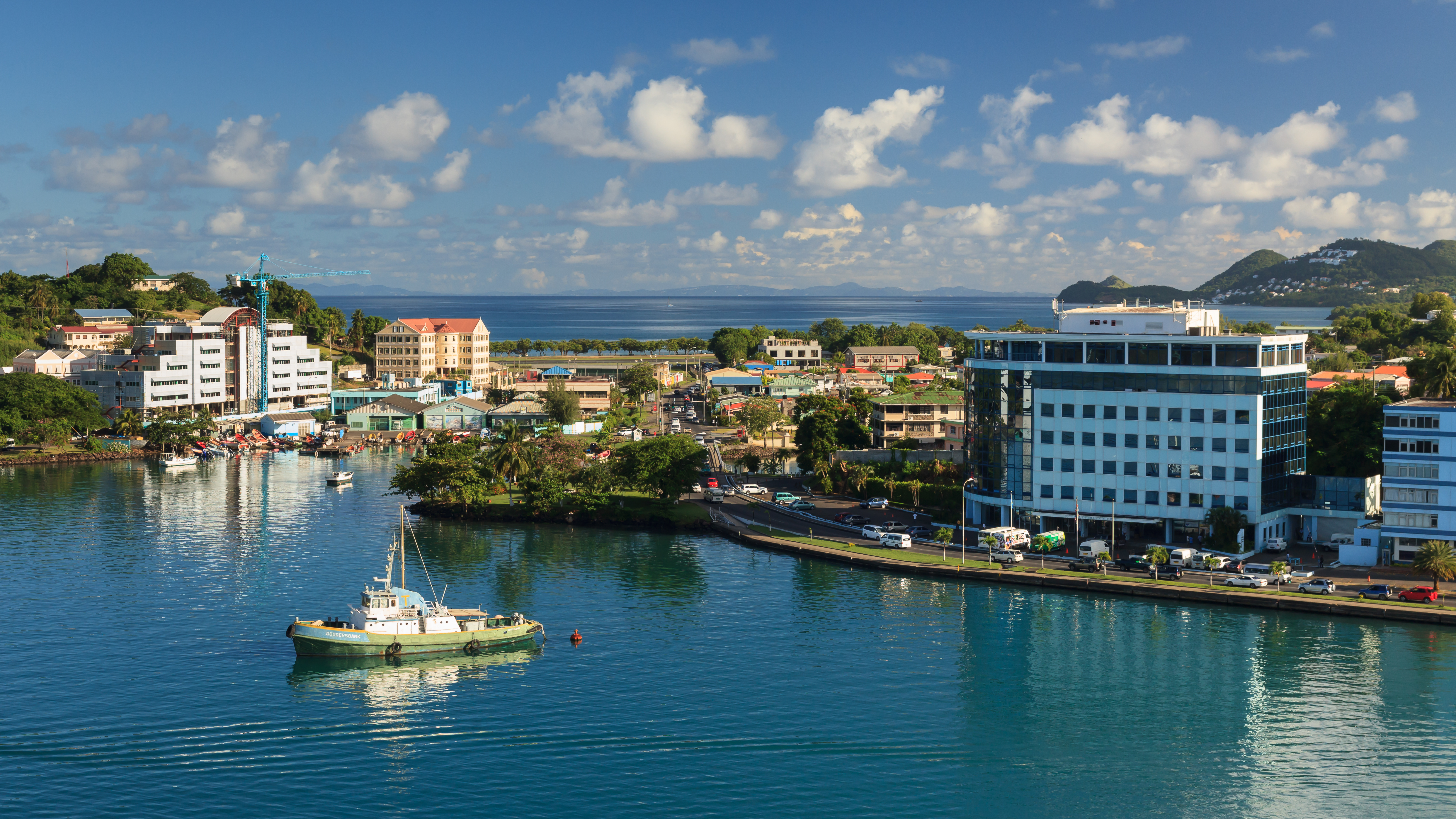 A scenic daytime view of Castries, St. Lucia, showing a calm blue harbor bordered by buildings, green trees, and distant hills under a partly cloudy sky. In the foreground, a light green and white boat floats on the water, while a coastal road with cars curves past a multi-story glass-and-concrete building on the right side of the bay.