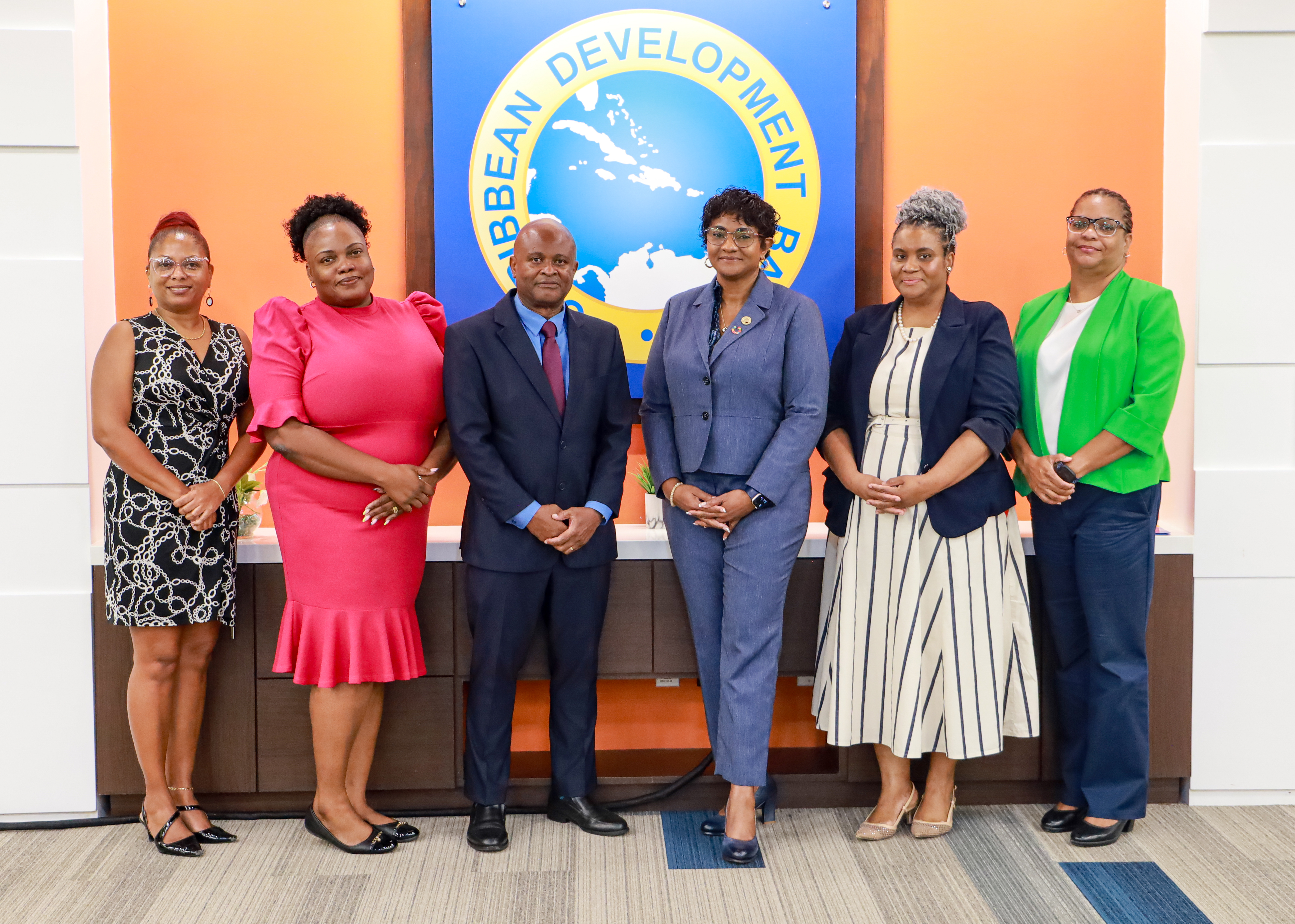 A group of six professionally dressed individuals—five women and one man—pose together indoors in front of a blue circular logo that reads "Caribbean Development Bank." The group stands in a single line against a backdrop featuring an orange accent wall and a dark wood counter. They are dressed in formal business attire, including colorful dresses, blazers, and a suit with a tie.
