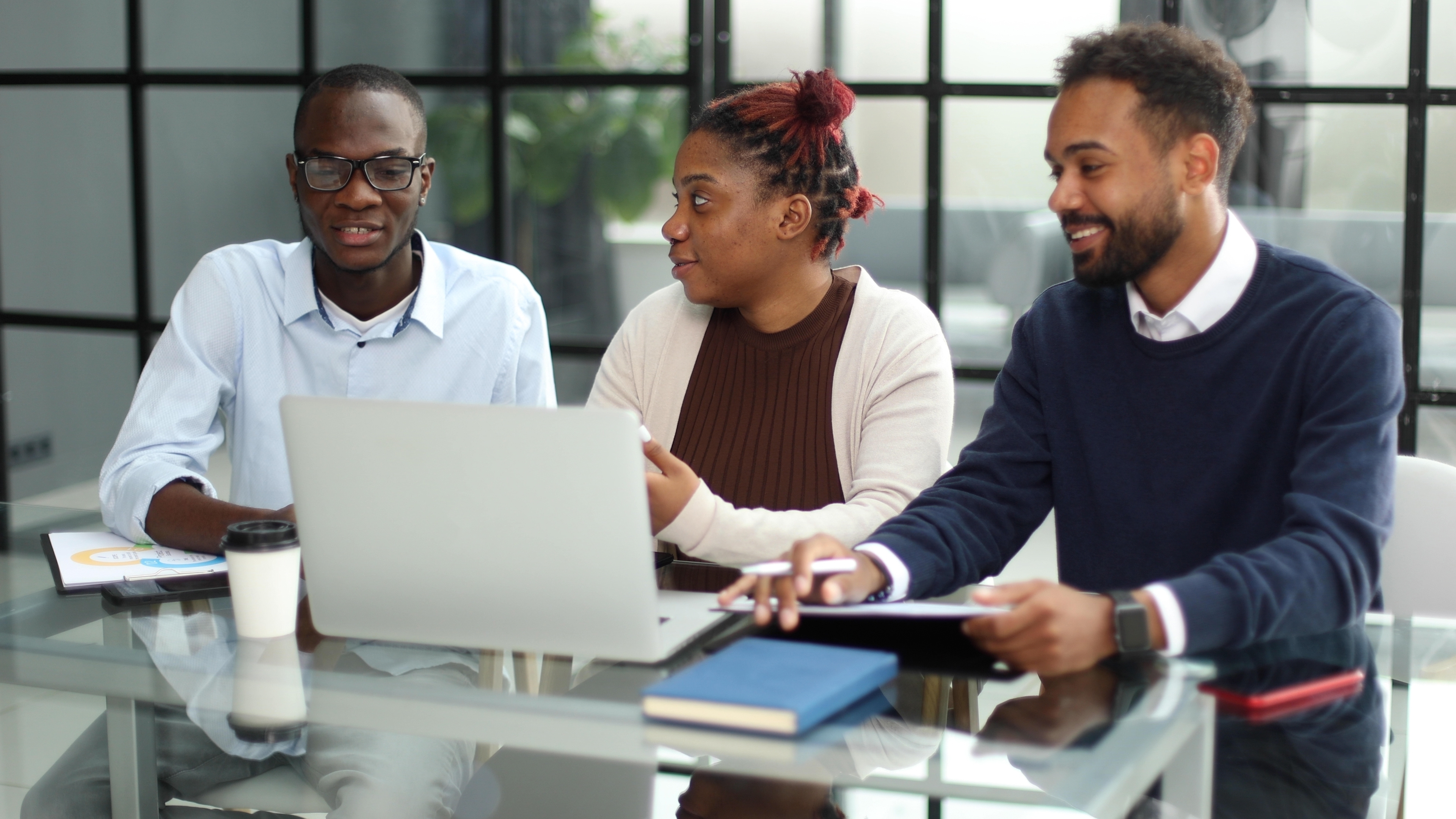 Three youth sitting in a boardroom in front of a laptop having a conversation. 