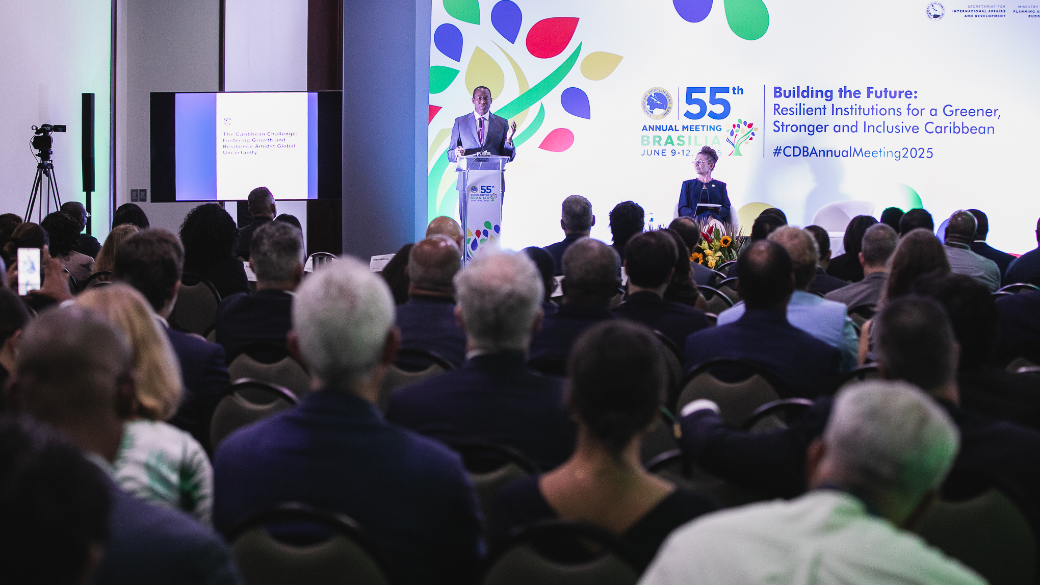 A large audience sits attentively in a conference room during the 55th Annual Meeting of the Caribbean Development Bank. Dr. Clarke stands at a lecte front, a speaern near a bright, colorful backdrop with the event’s branding and theme. The scene captures an engaged crowd focused on the presentation.