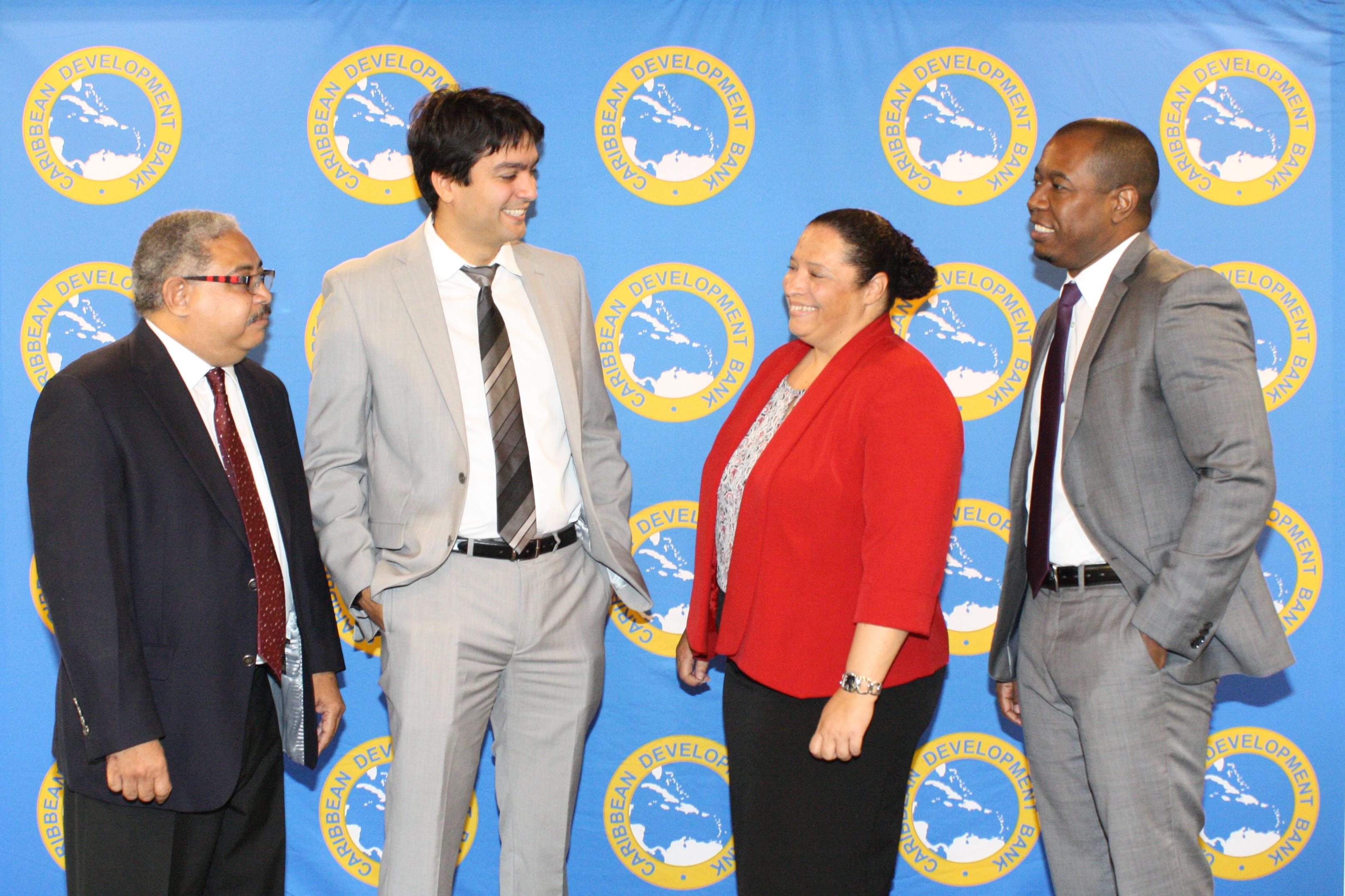 Four people in suits talk with each other, smiling, against a backdrop featuring CDB's logo