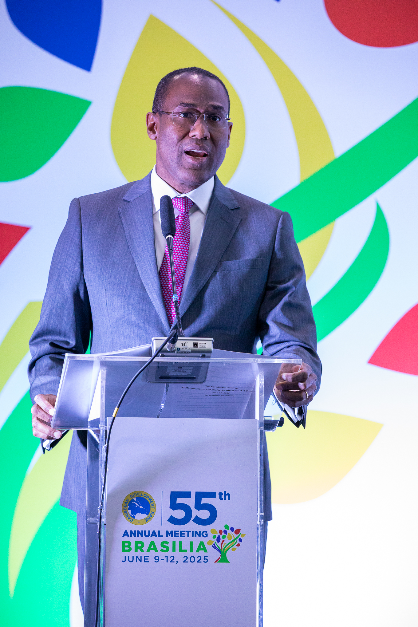 Dr. Clarke in a gray suit stands behind a clear lectern, delivering a speech at the 55th Annual Meeting of the Caribbean Development Bank in Brasília, Brazil. The background features colorful graphics and the event's logo, emphasizing themes of resilience, inclusivity, and sustainability.
