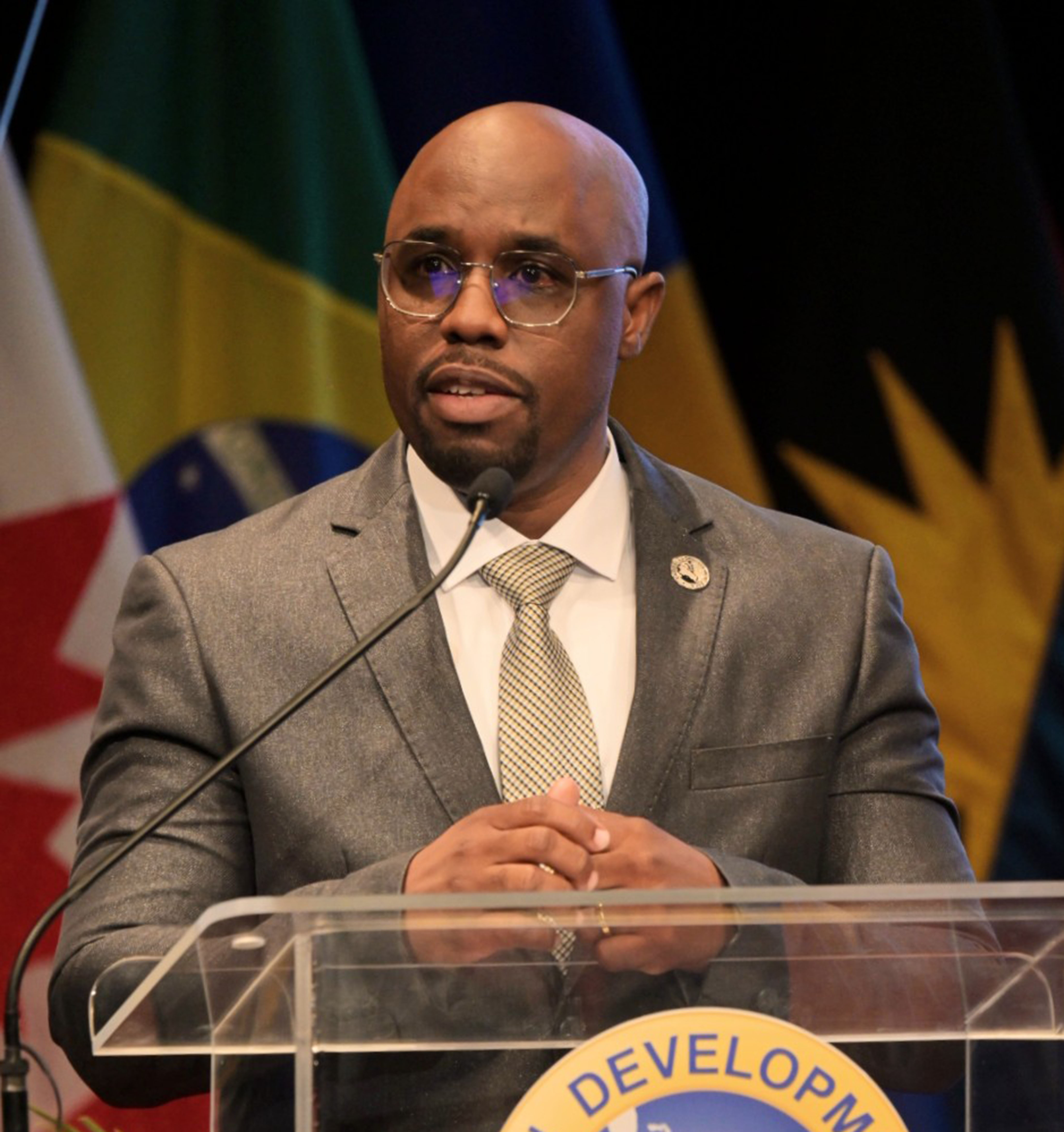 Speaker in a grey suit standing at a clear podium with Caribbean flags displayed behind.