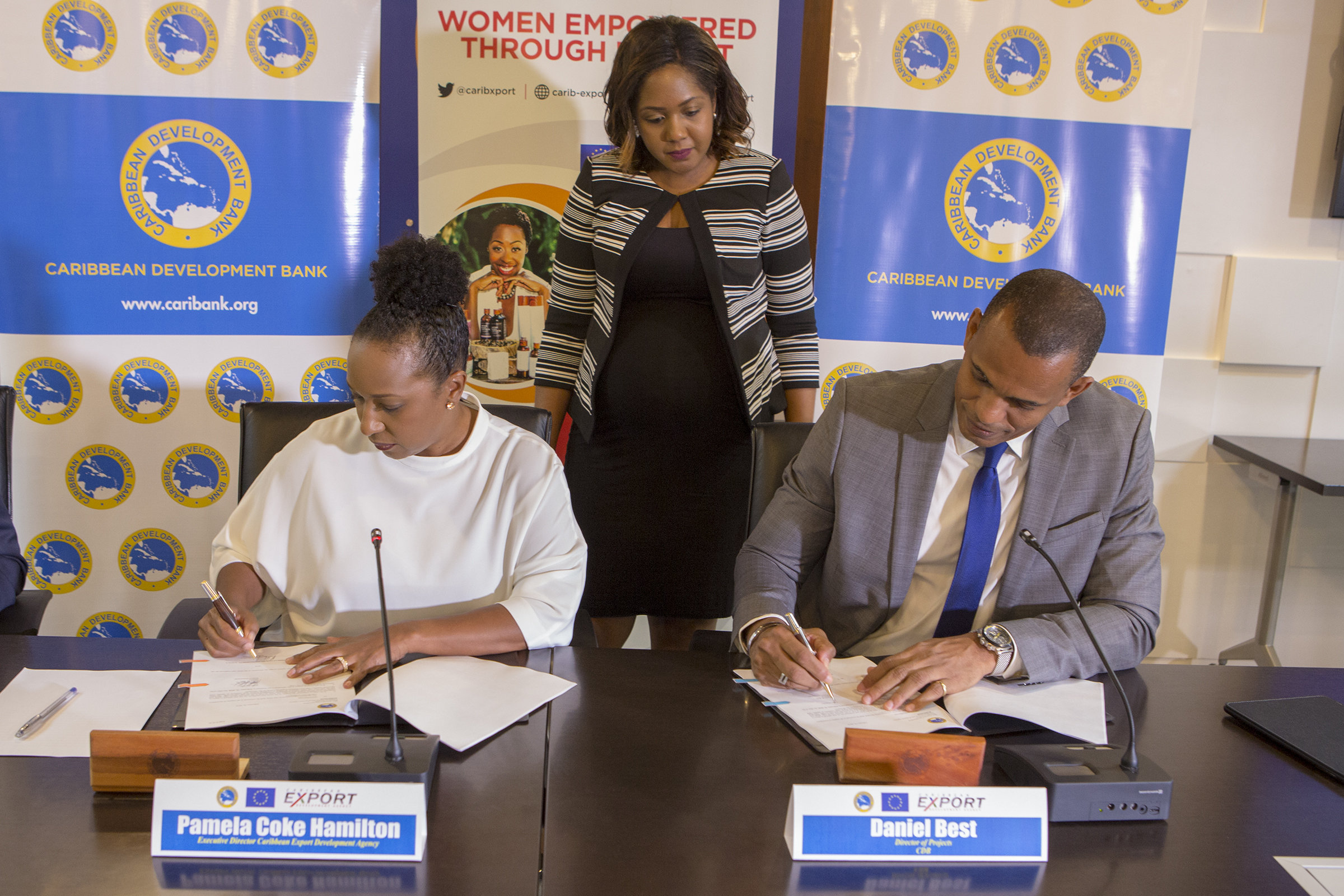 Pamela Coke Hamilton, Executive Director, Caribbean Export (left) and Daniel Best, Director of Projects, CDB (right) sign the WE-Xport Agreement, while Alana Goodman-Smith, Legal Counsel, CDB (standing) observes.