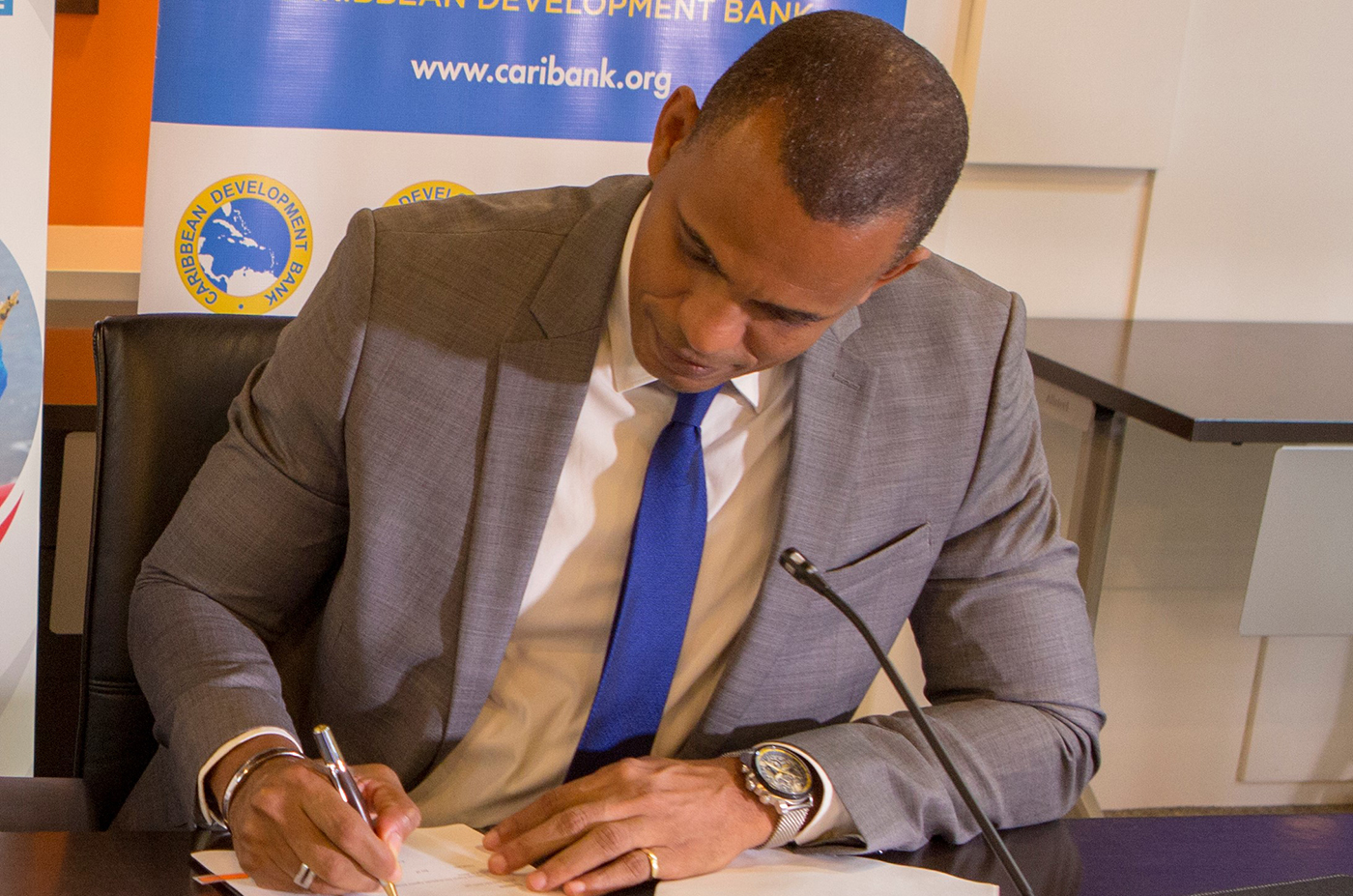 Daniel Best wearing grey suit with blue tie signing document.