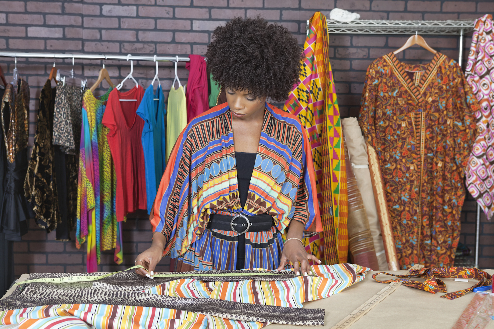Female fashion designer measuring fabric surrounded by colourful outfits